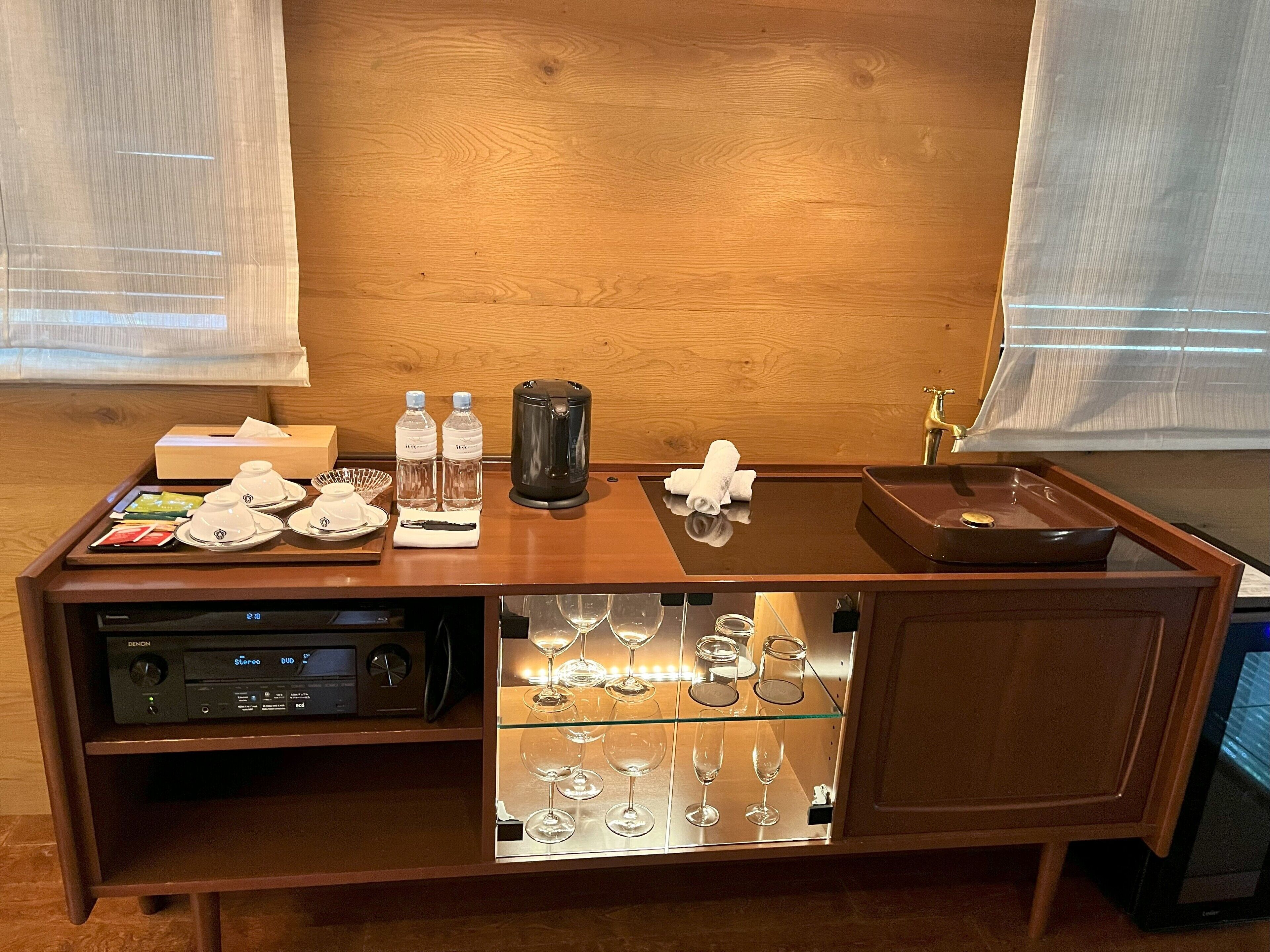 A wooden bar area with a coffee maker, water bottles, and a glass display case with wine glasses.