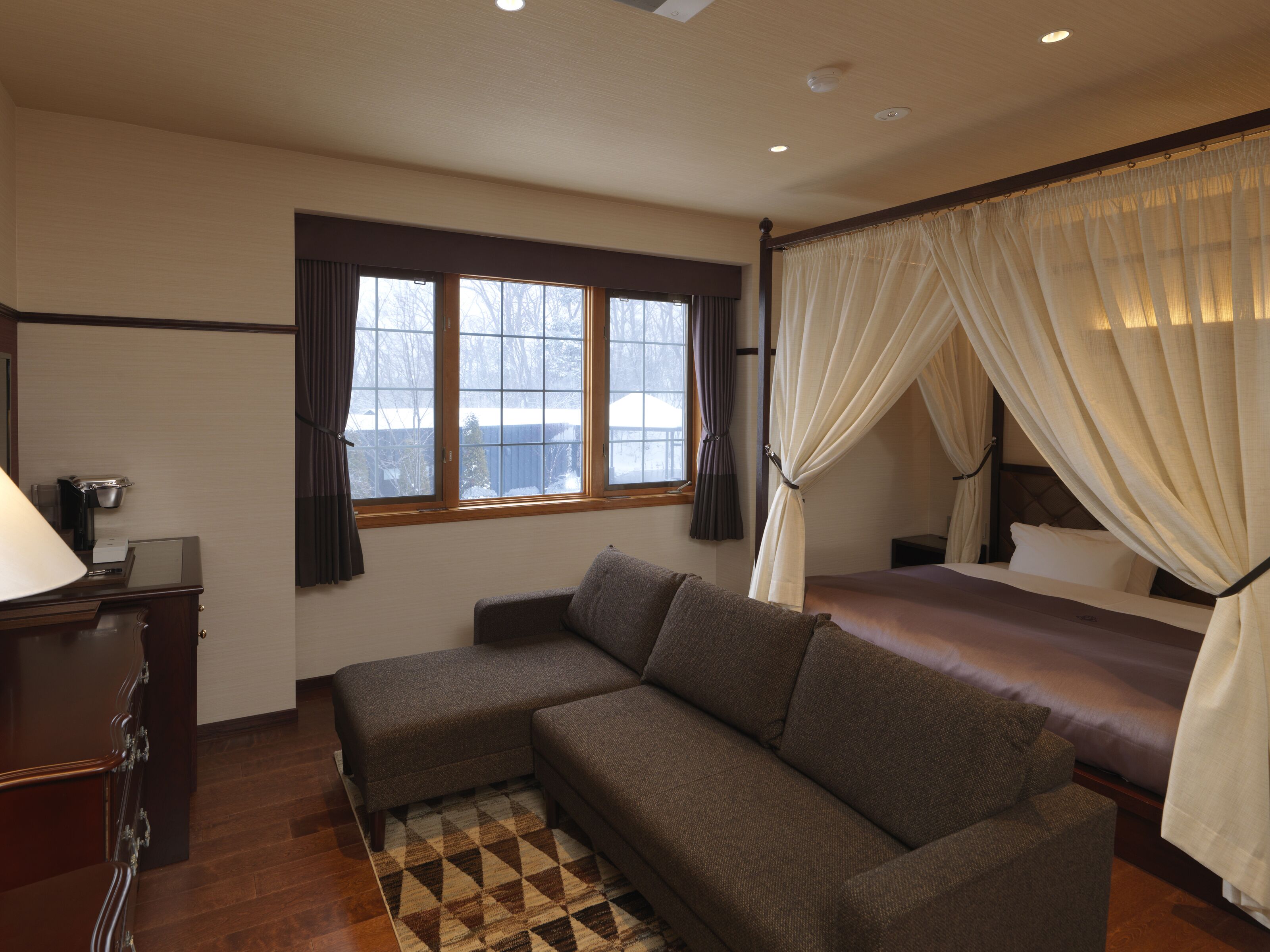 A bedroom with a sofa, a bed with a canopy, a desk with a lamp, and a window with curtains.