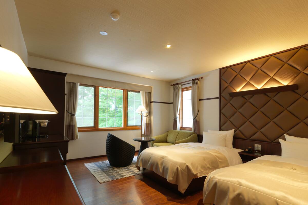 A hotel room with two beds, a desk with a lamp, a window with curtains, and a wall with a textured panel.