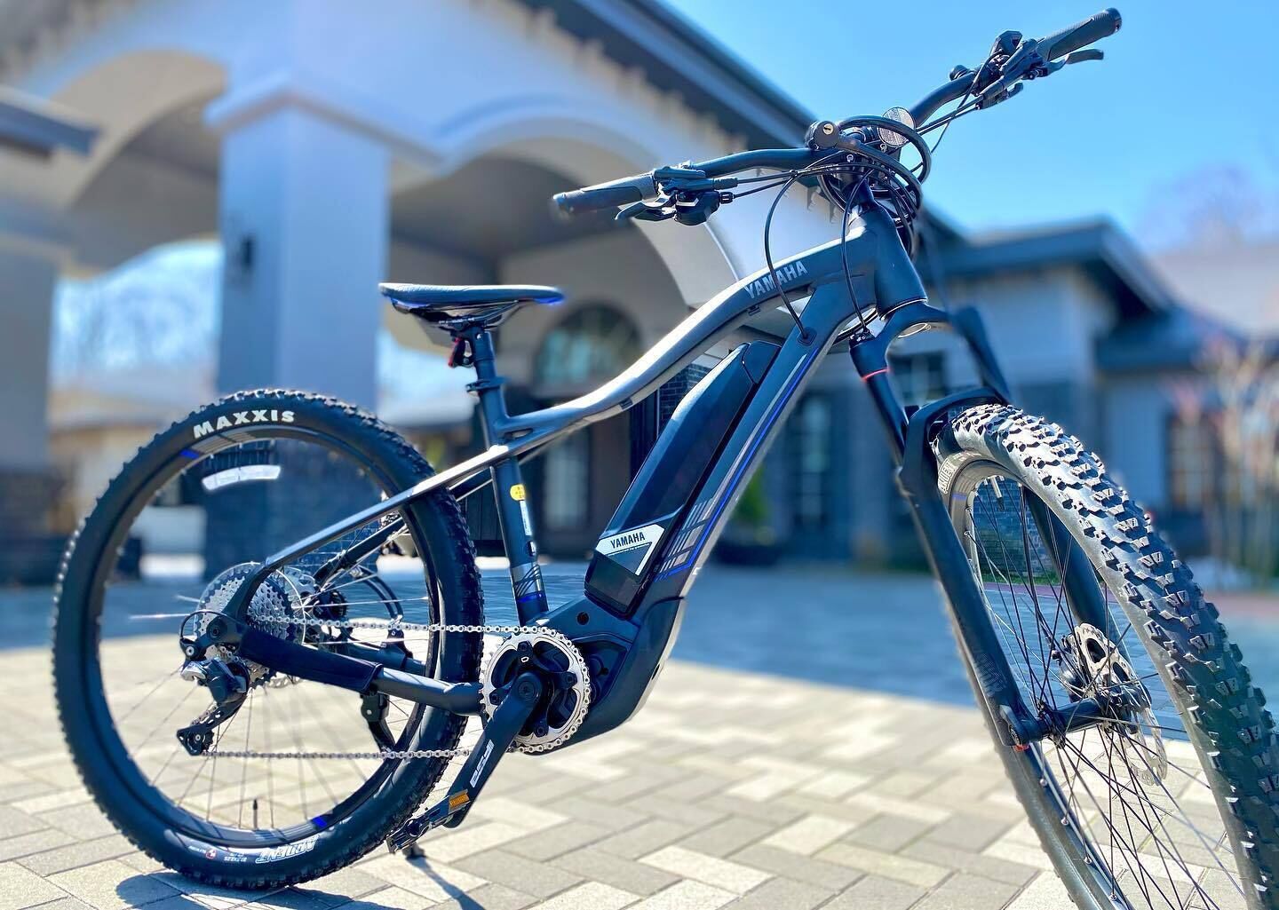 A blue electric bicycle with a rear rack, a chain, and a Shimano gear system, parked on a paved surface.