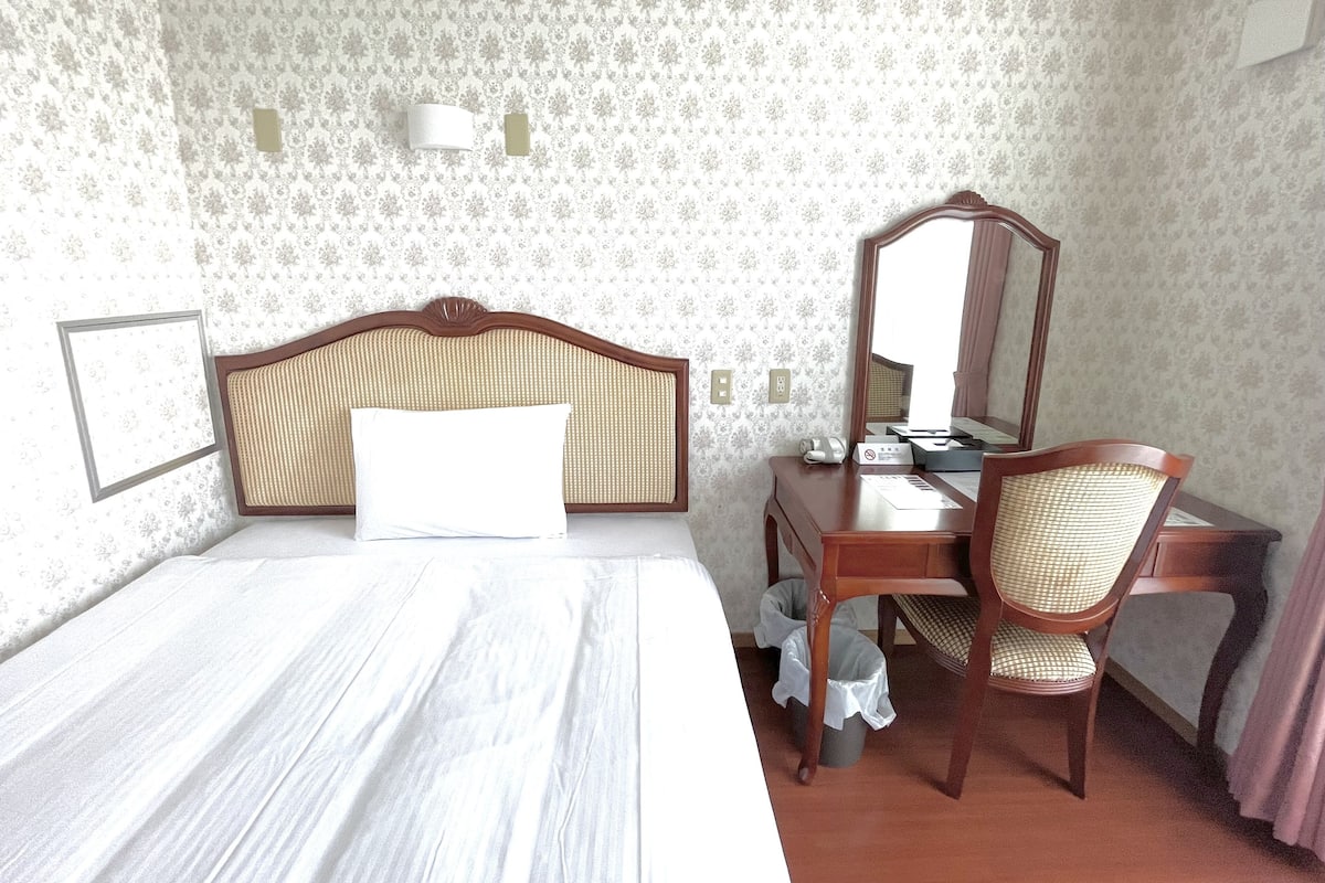 A hotel room with a bed, a desk, a chair, a mirror, and a wall-mounted light fixture.