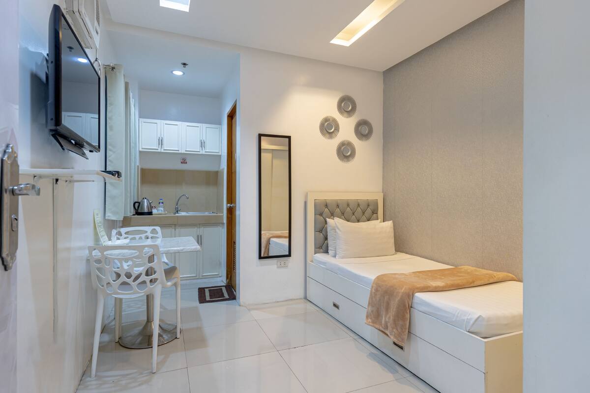 A compact bedroom with a bed, a bedside table, and a chair. There is a small kitchenette with a sink and cabinets.