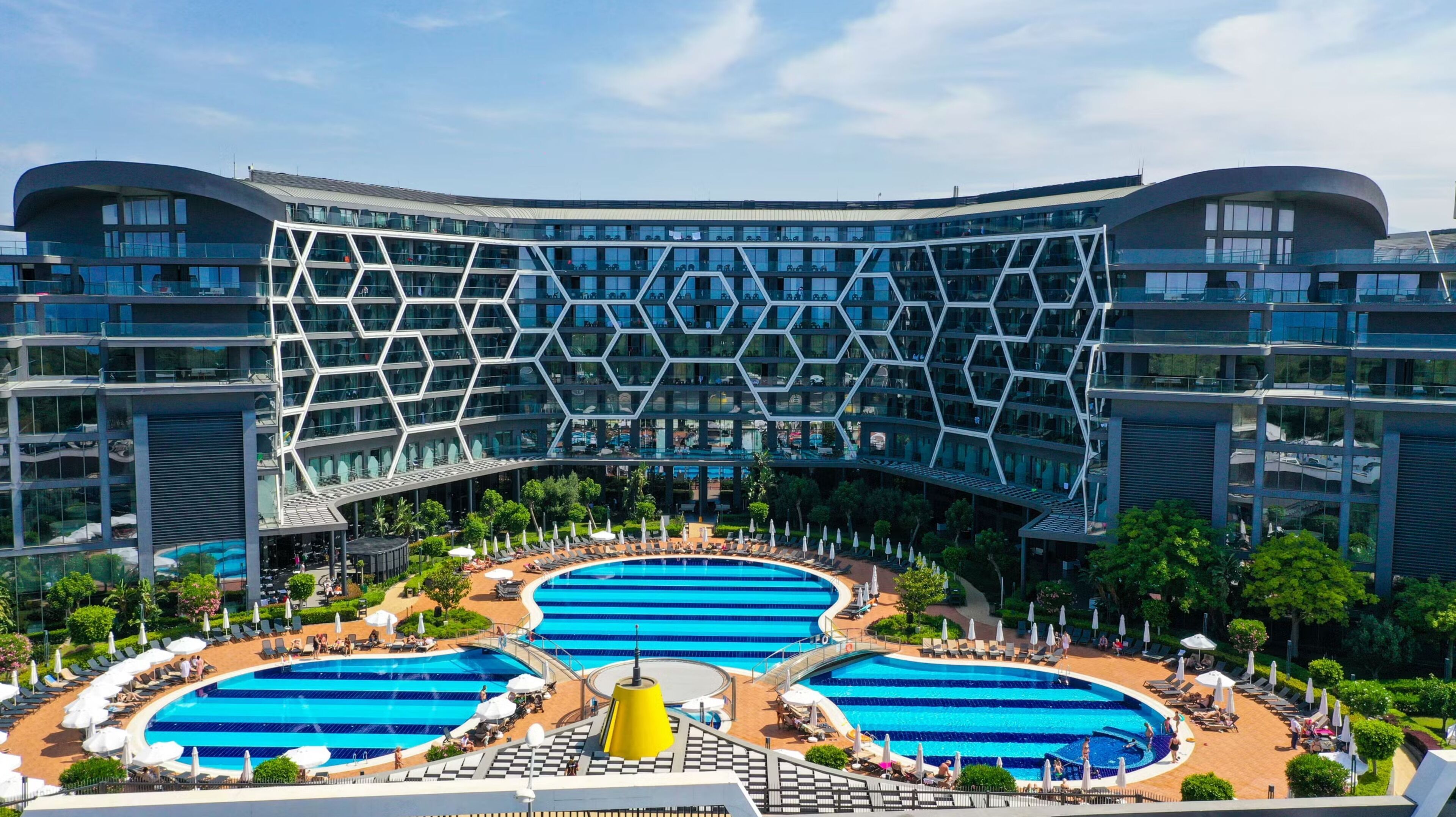 Un hotel moderno con una fachada geométrica distintiva, múltiples piscinas y un área exterior bien cuidada con tumbonas y sombrillas.