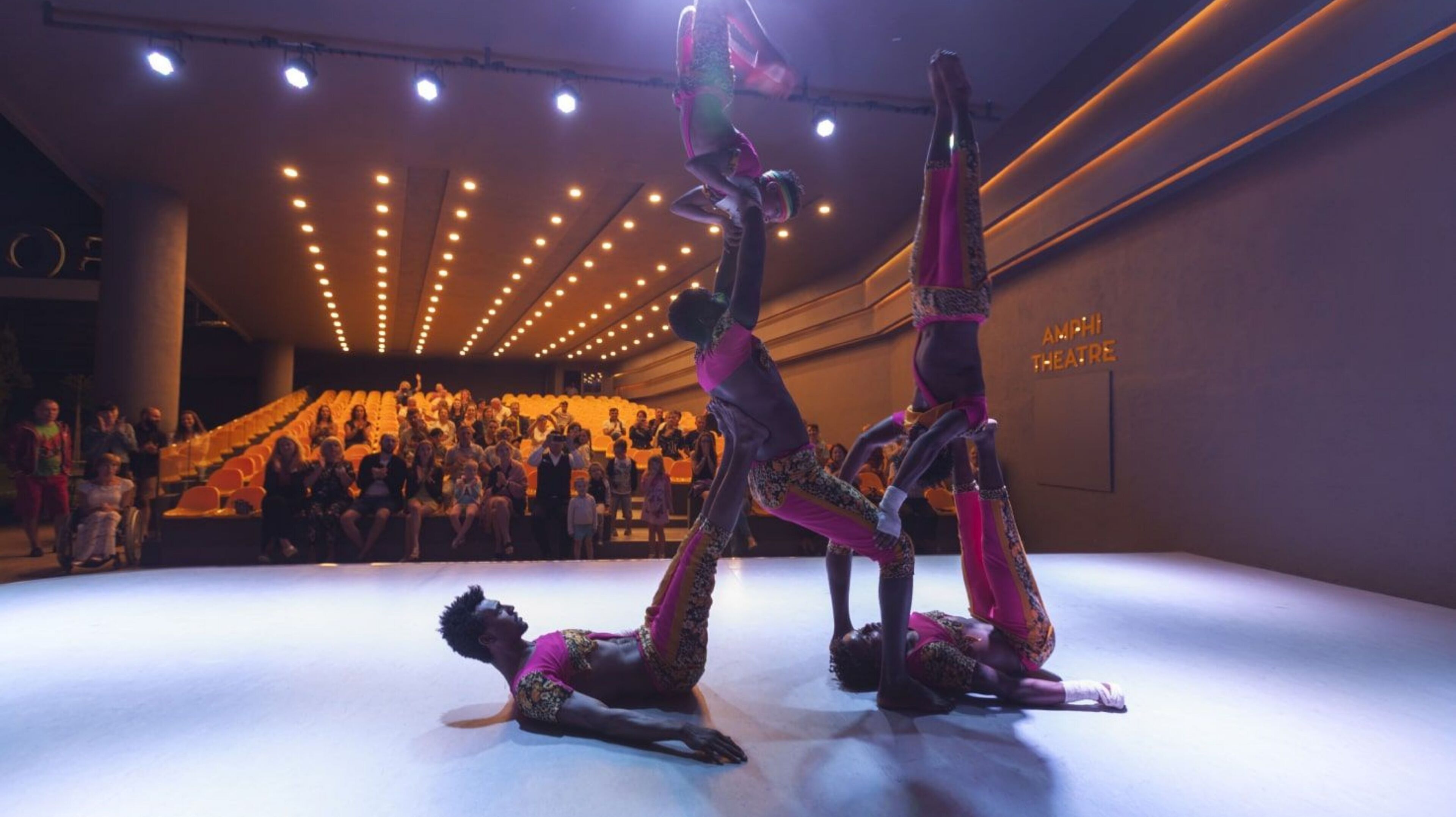 Een optreden in een zaal met twee acrobaten in de lucht, een zittend publiek en een podium met decor.
