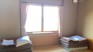 A room with tatami mats, two low beds with pillows, a window with curtains, and a wall-mounted air conditioning unit. - Hakodateyama Guest House - Hostel (Hakodate)