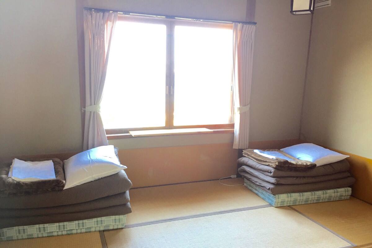 A room with tatami mats, two low beds with pillows, a window with curtains, and a wall-mounted air conditioning unit.