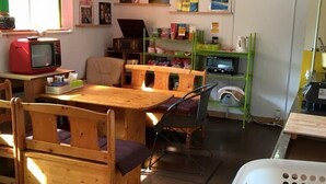 A room with a wooden dining table, chairs, a television, a bookshelf, and a washing machine. - Hakodateyama Guest House - Hostel (Hakodate)