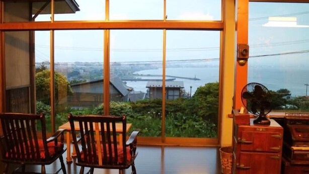 A room with large windows offering a view of a body of water and buildings. There are two wooden chairs and a small table with a fan. - Hakodateyama Guest House - Hostel (Hakodate)