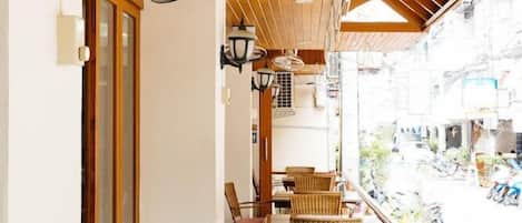 An indoor seating area with wicker chairs and tables, wooden ceiling beams, and wall-mounted lamps.