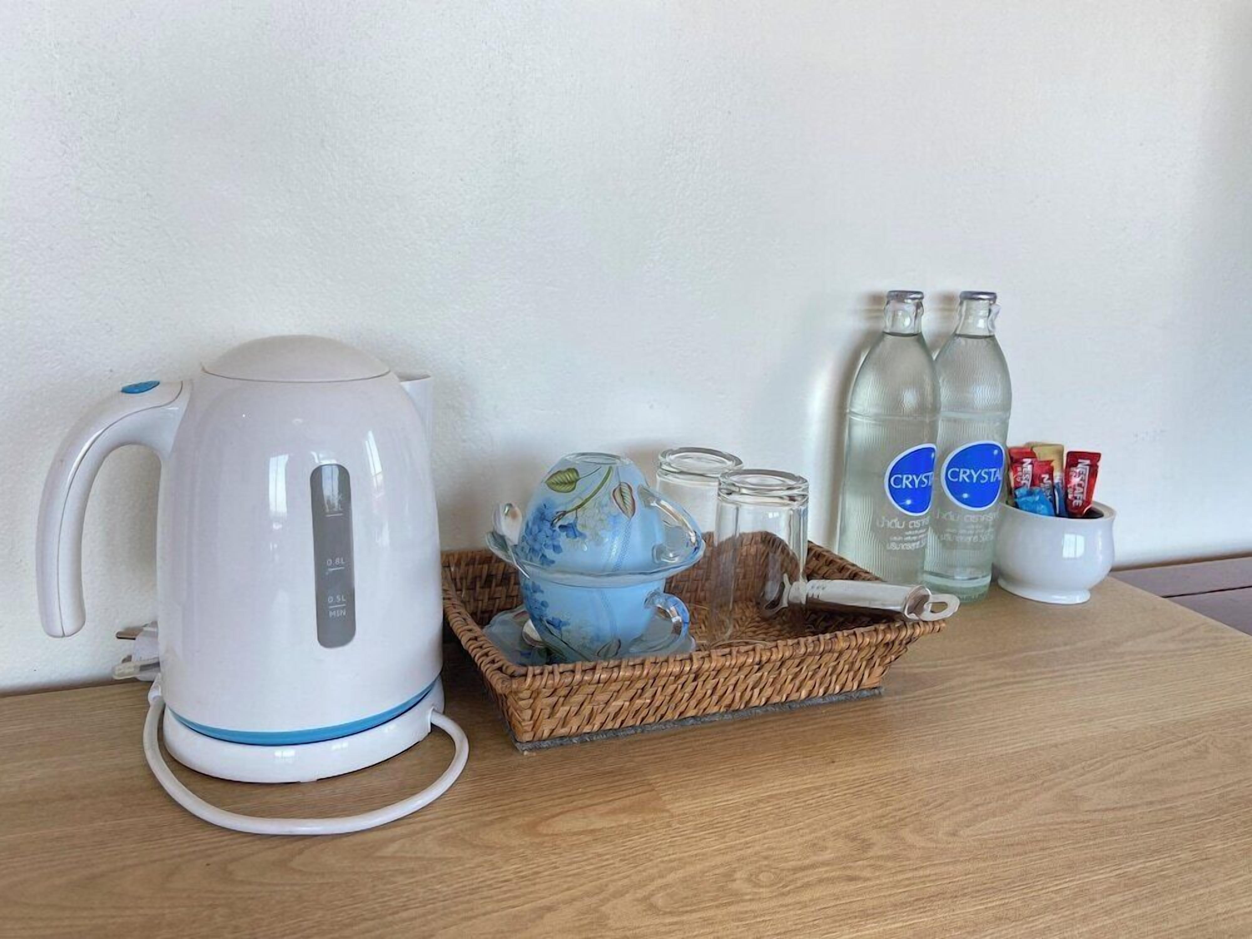 Une bouilloire électrique blanche, deux bouteilles d’eau, une théière en verre et un panier contenant des tasses et des en-cas, le tout posé sur une table en bois.