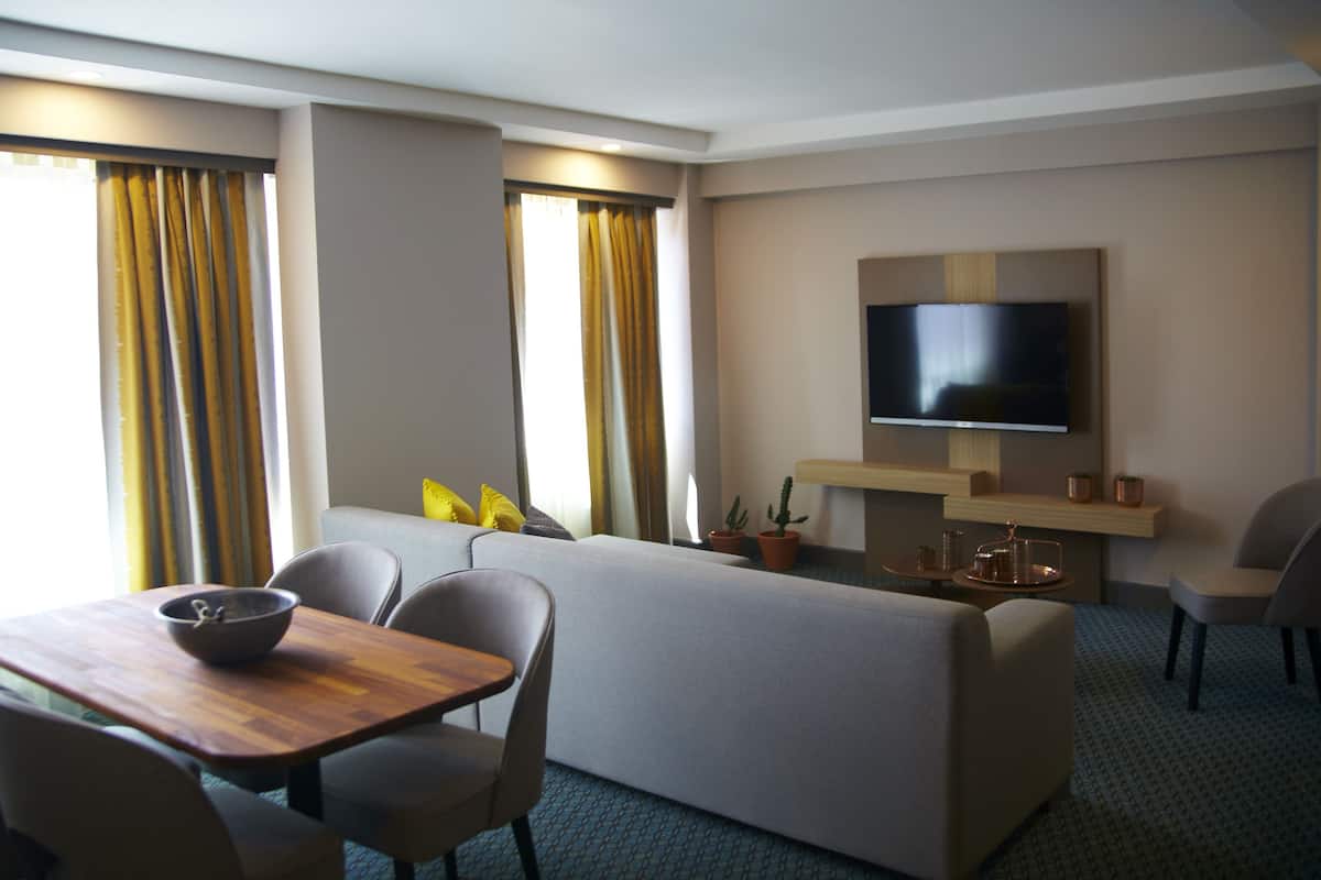 A hotel room with a sofa, a TV mounted on the wall, a dining table with chairs, and a small table with a bowl.