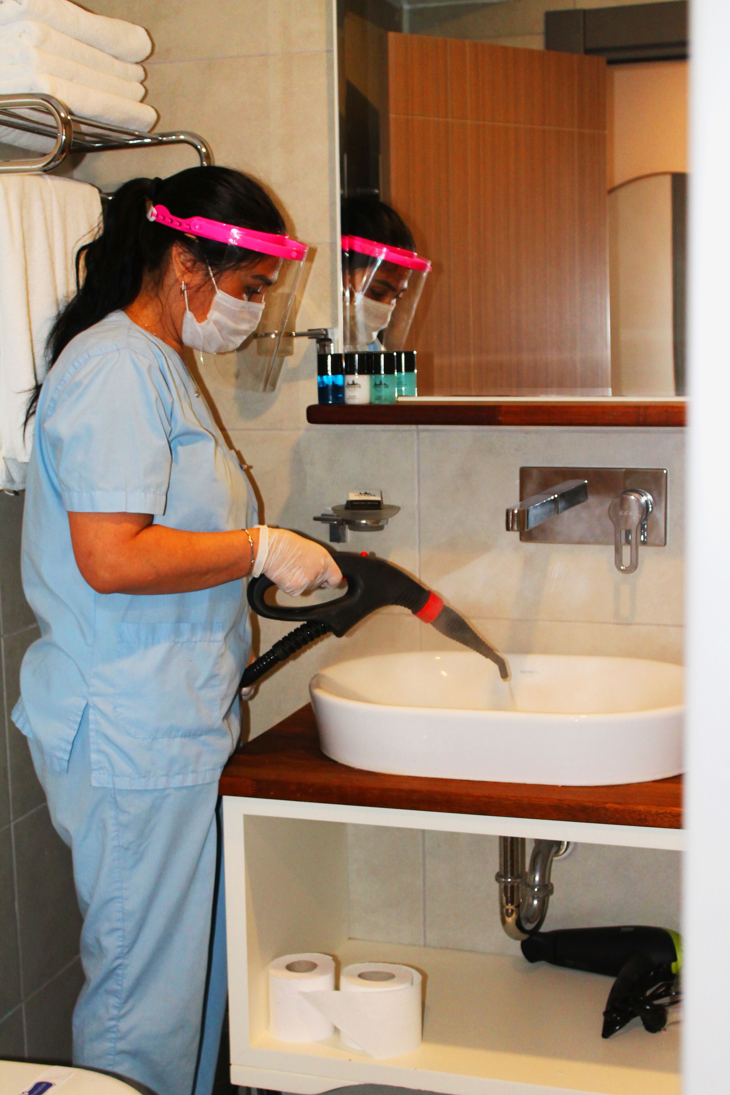 A person in a blue uniform cleaning a sink with a high-pressure water hose in a bathroom with a wooden vanity and a mirror above the sink.