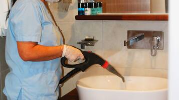 A person in a blue uniform cleaning a sink with a high-pressure water hose in a bathroom with a wooden vanity and a mirror above the sink.