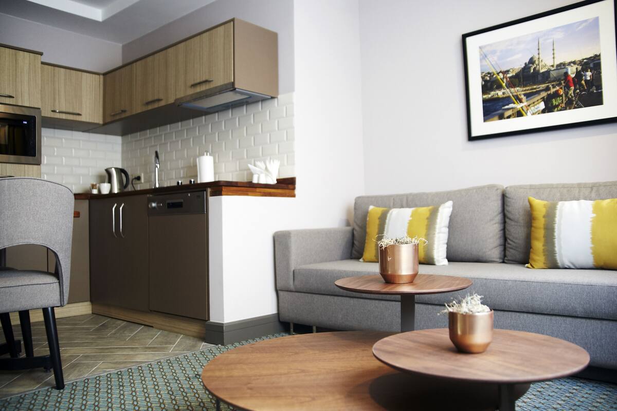 A modern kitchen with wooden cabinets, a grey sofa with yellow and white cushions, and a round wooden coffee table.