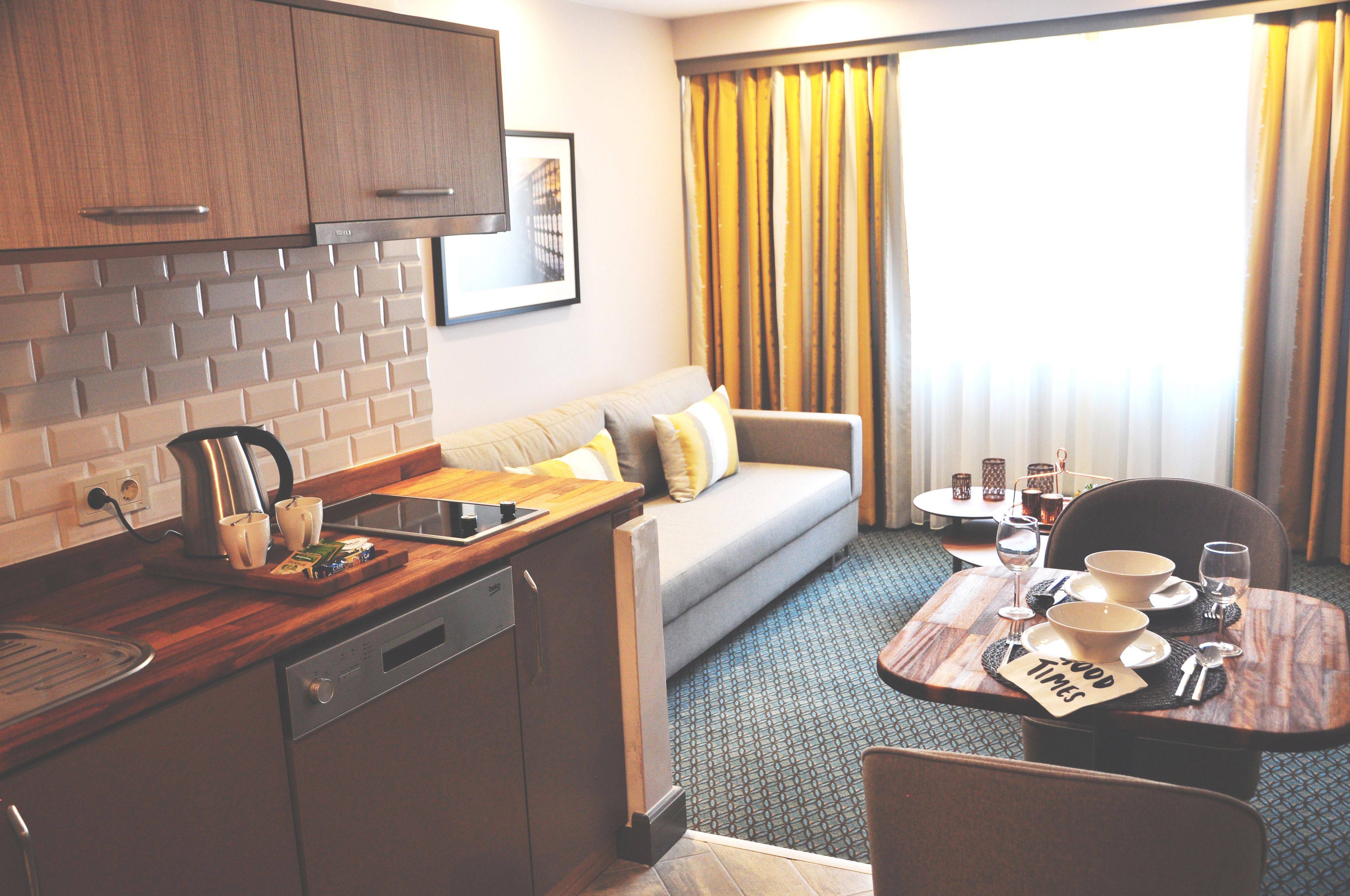 Habitación de hotel compacta con kitchenette, sofá, mesa de comedor con desayuno listo y un amplio ventanal con cortinas.