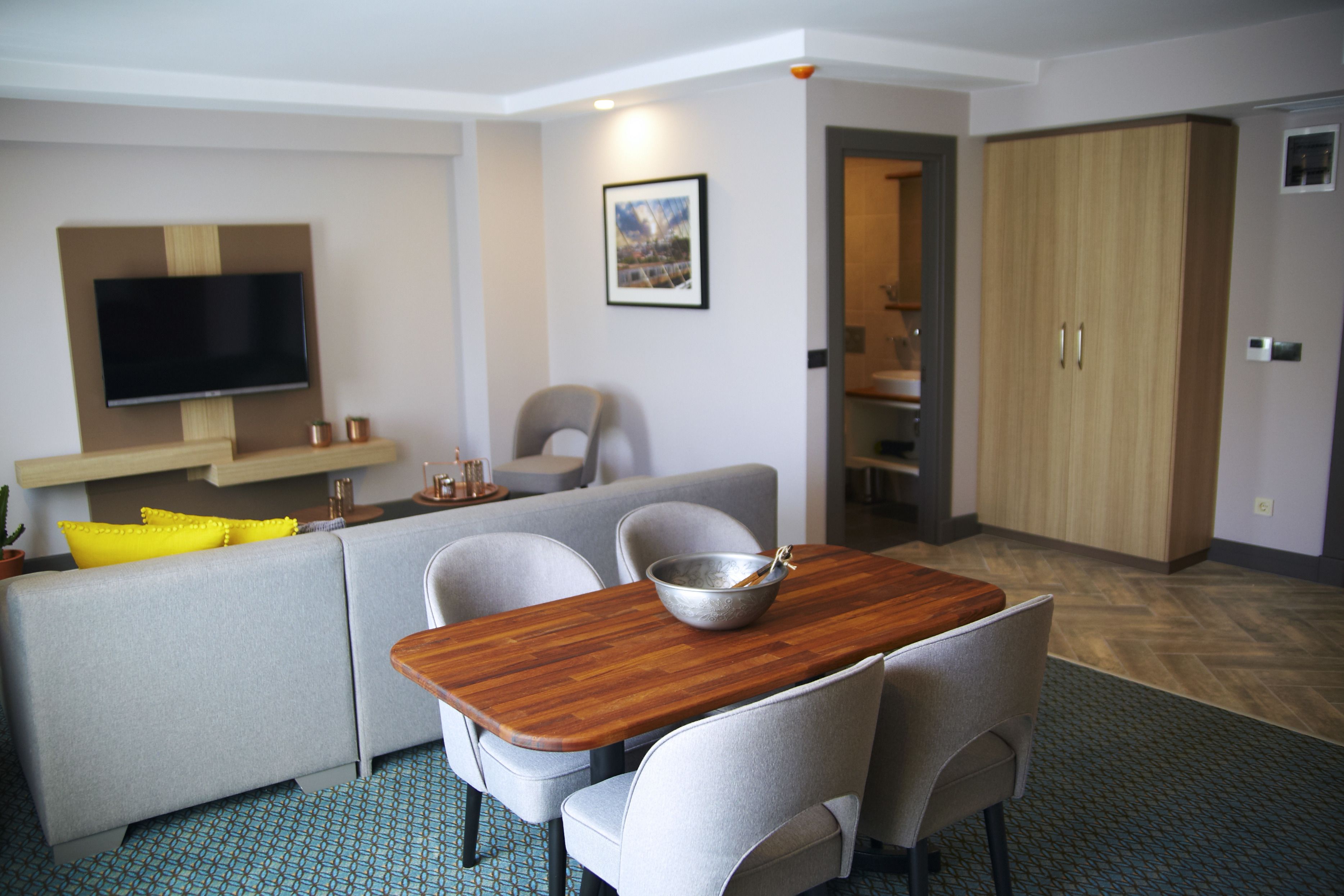 A modern hotel room with a dining area, a sofa, a TV, and a wardrobe.