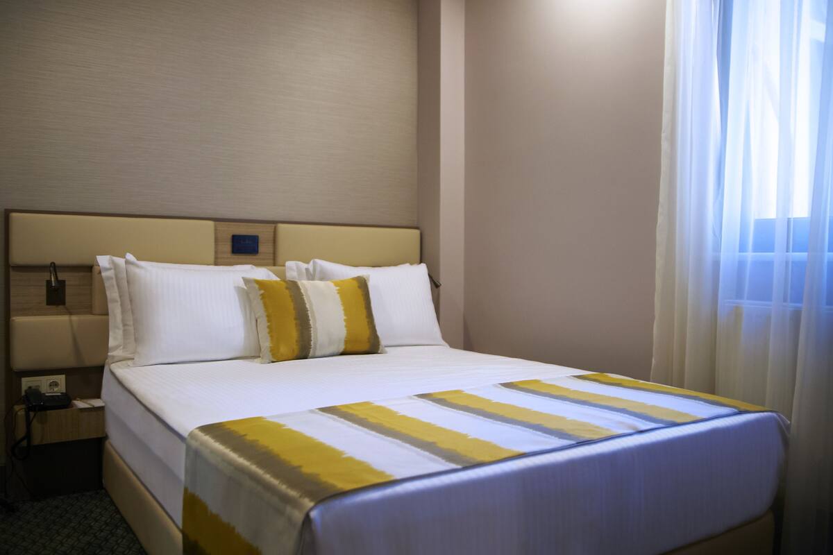 A hotel room with a bed, a nightstand, a lamp, and a window with curtains.