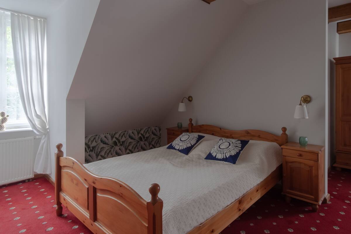 Un dormitorio con techo inclinado, una cama de madera con dos almohadas decorativas, una mesita de noche con lámpara y una ventana con cortinas.