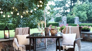 Un patio al aire libre con sillas de mimbre, una mesa de vidrio, velas y luces colgantes.
