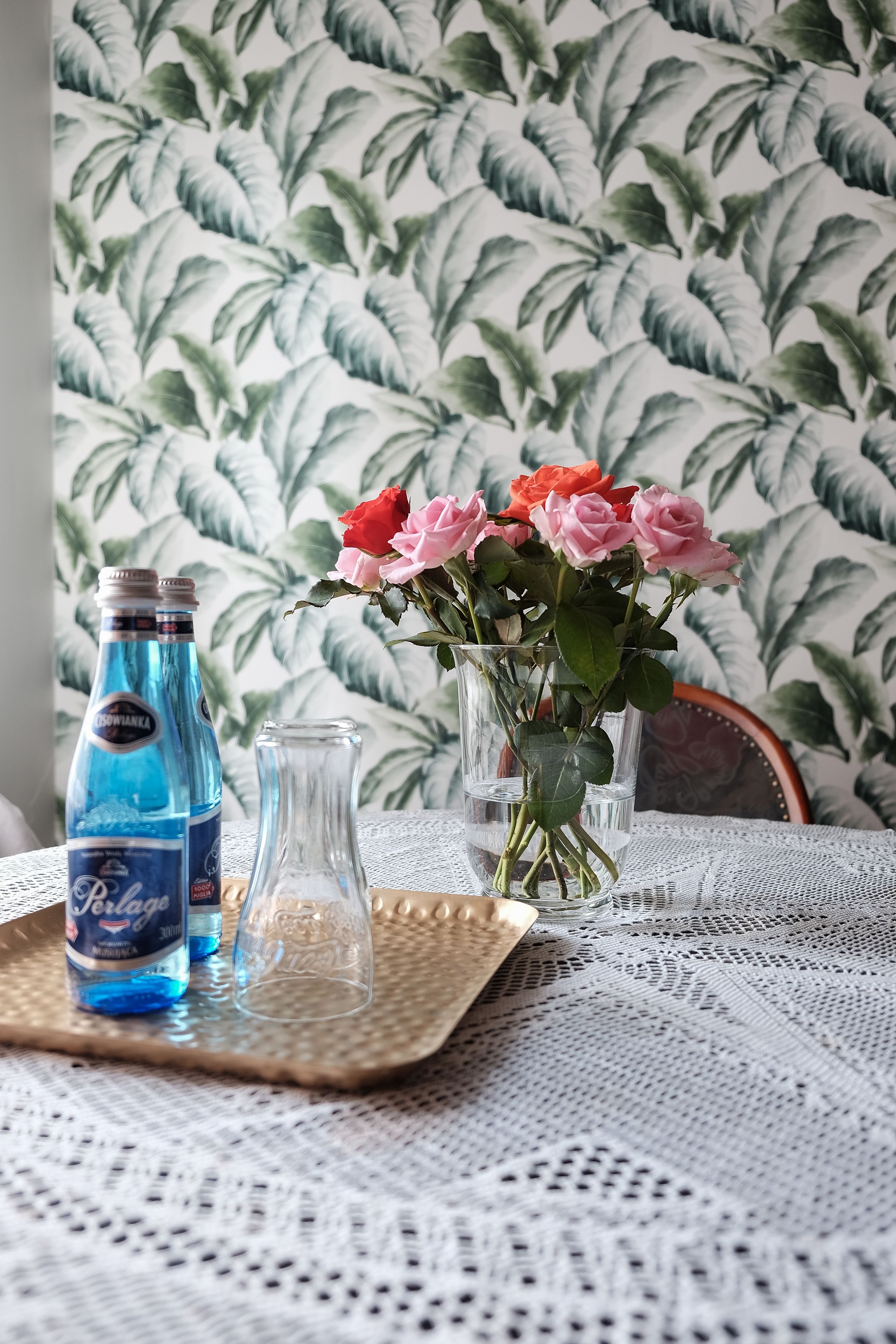 Une table avec une bouteille d’eau, un vase en verre contenant des roses roses et un verre transparent.