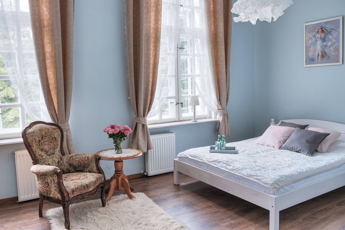 Un dormitorio con cama, una silla, una mesita con un jarrón de flores y una ventana con cortinas.