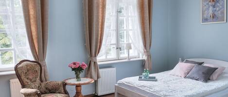 Un dormitorio con cama, una silla, una mesita con un jarrón de flores y una ventana con cortinas.