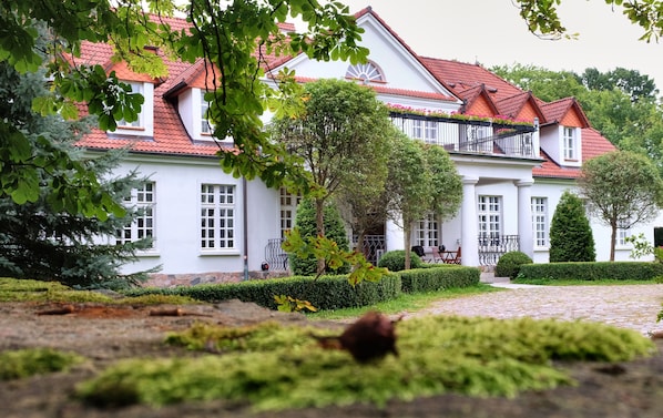 A large white building with a red-tiled roof, surrounded by greenery and a cobblestone path.