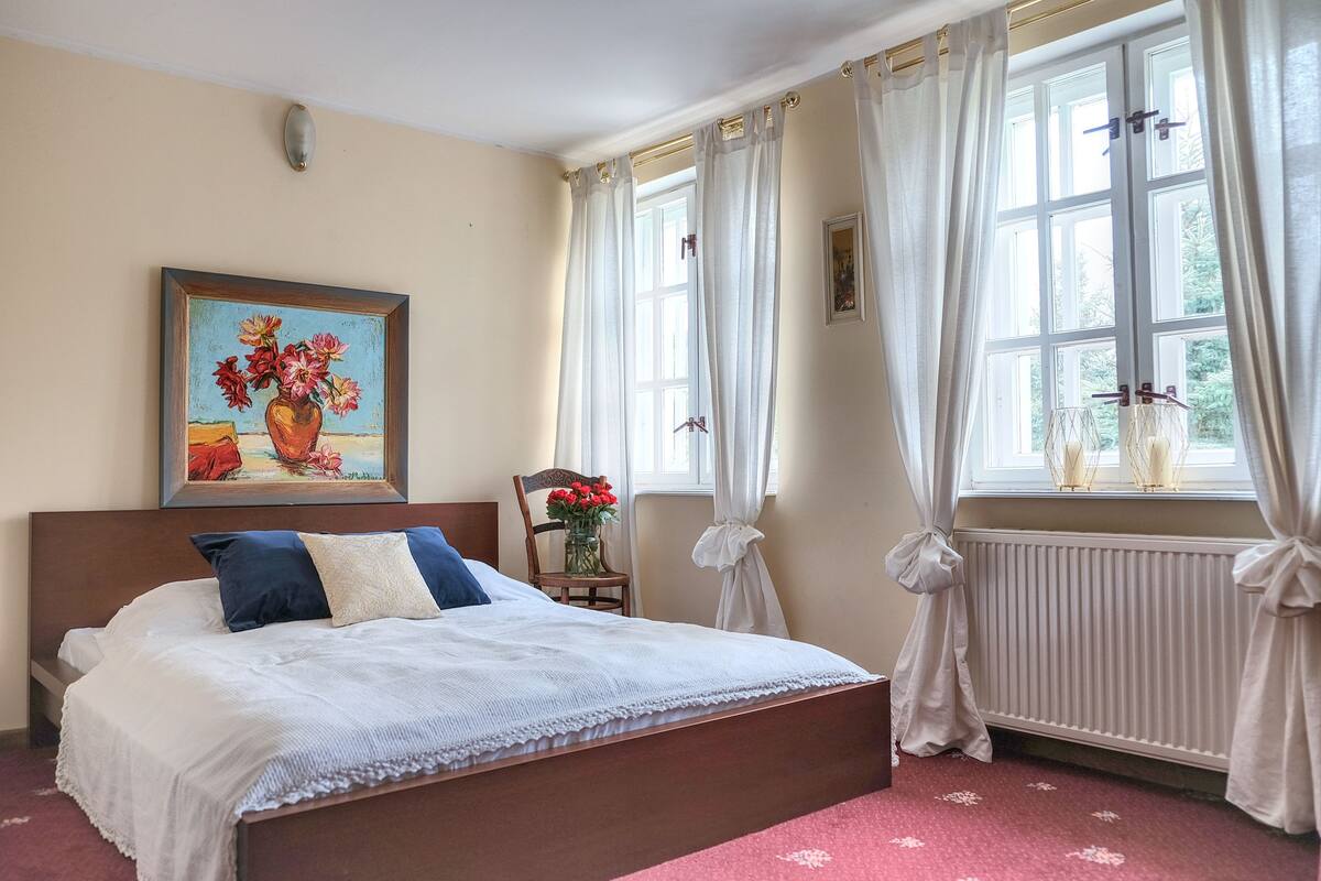 Un dormitorio con cama, un cuadro, una silla con un florero y una ventana con cortinas.
