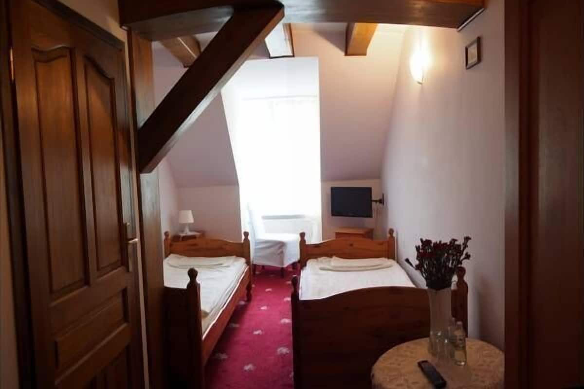 Un pasillo estrecho que da a un dormitorio con dos camas individuales, una mesita con un florero y un televisor en la pared.