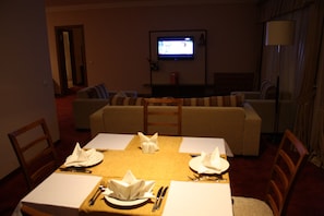 A dining area with a table set for four, a television mounted on the wall, and a sofa in the background.