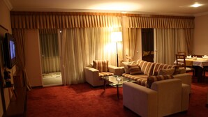A hotel room with a red carpet, beige sofa, armchair, and a glass coffee table. There is a TV on the left and a dining table with chairs on the right. - Qafqaz Sahil Resort Hotel (Lankaran)