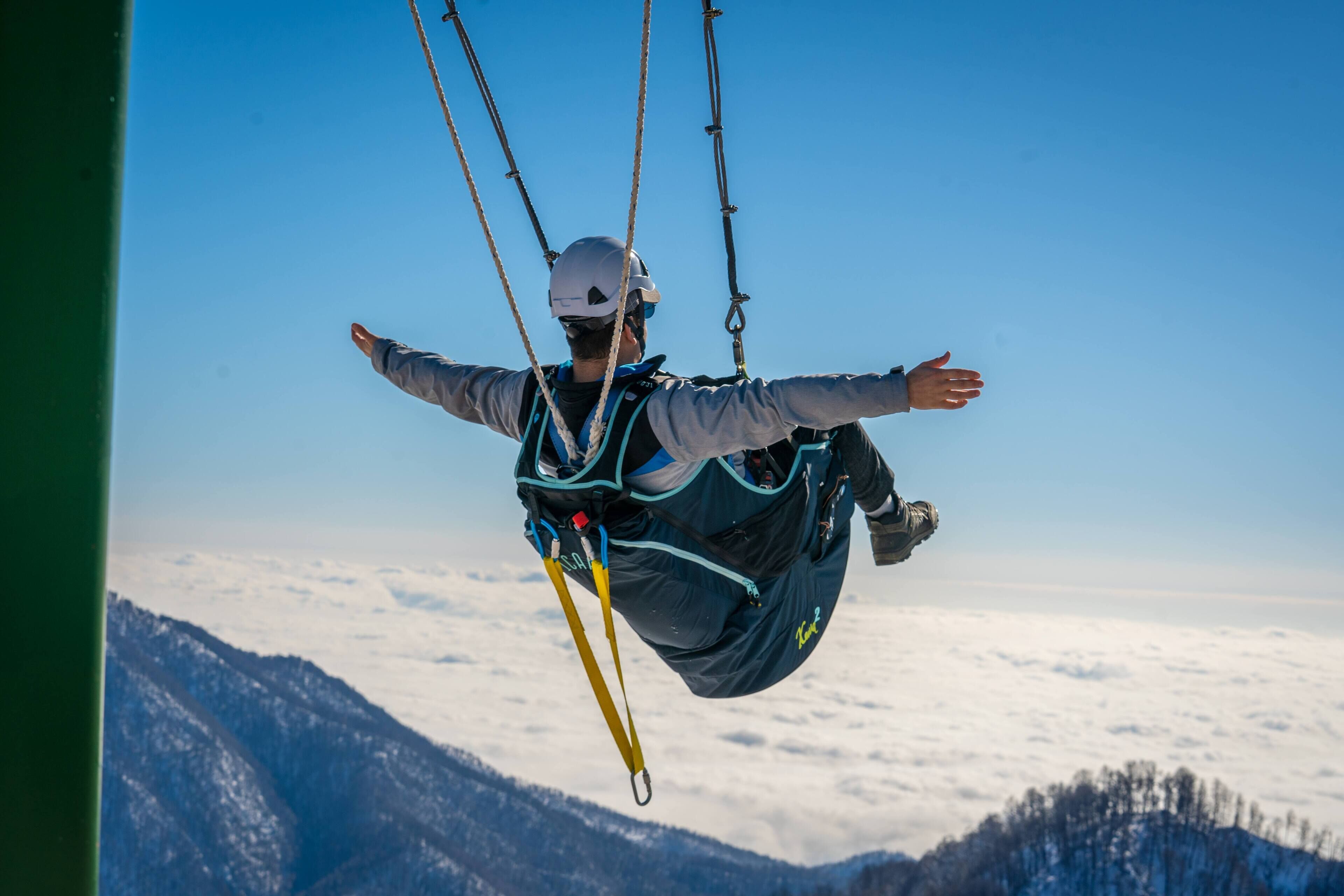 Une personne pratiquant le parachutisme, les bras tendus, avec un harnais et un casque, sur fond de montagnes et de nuages.