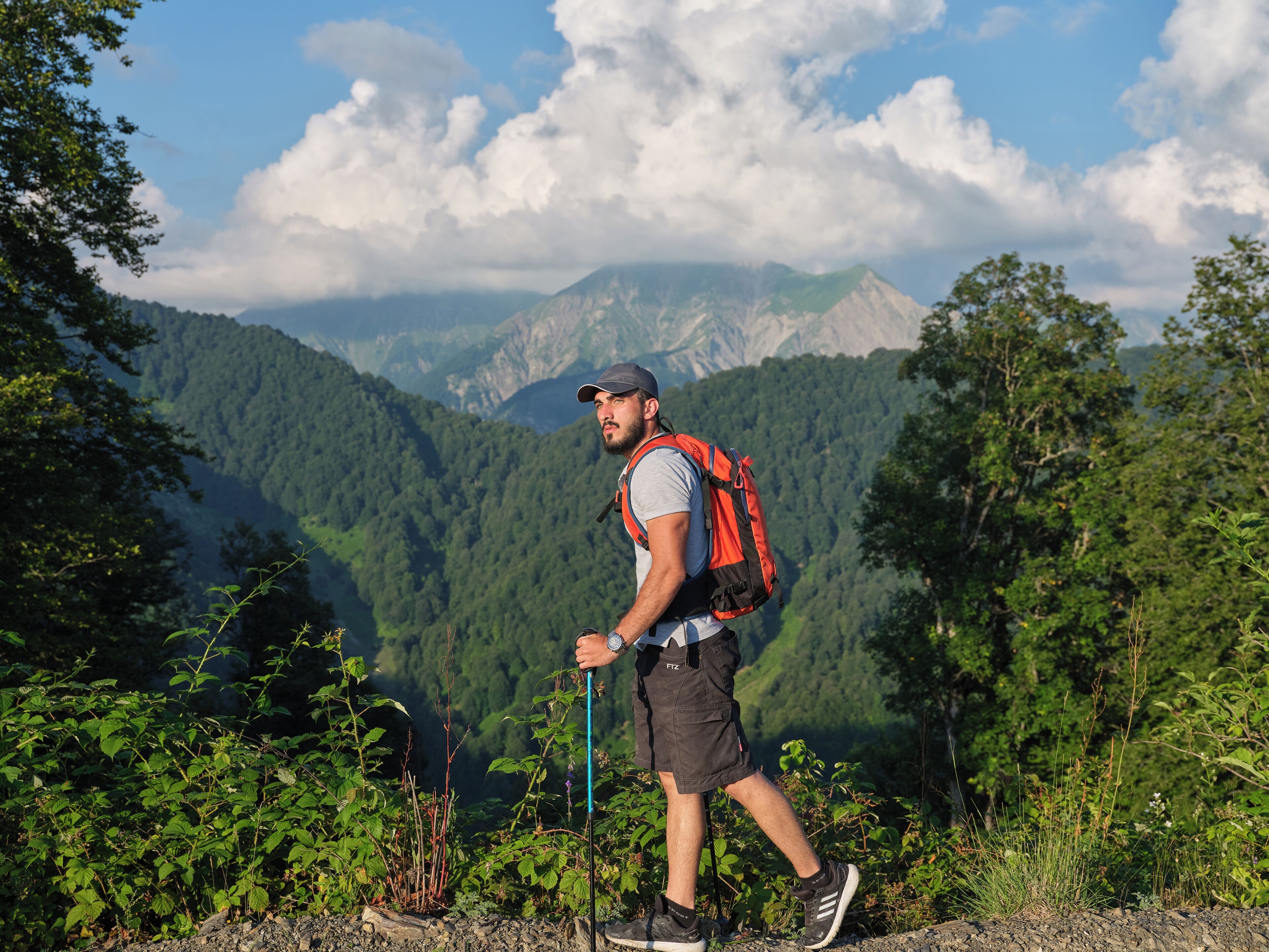 A hiker with a backpack and walking stick on a rocky path with greenery and mountains in the background.