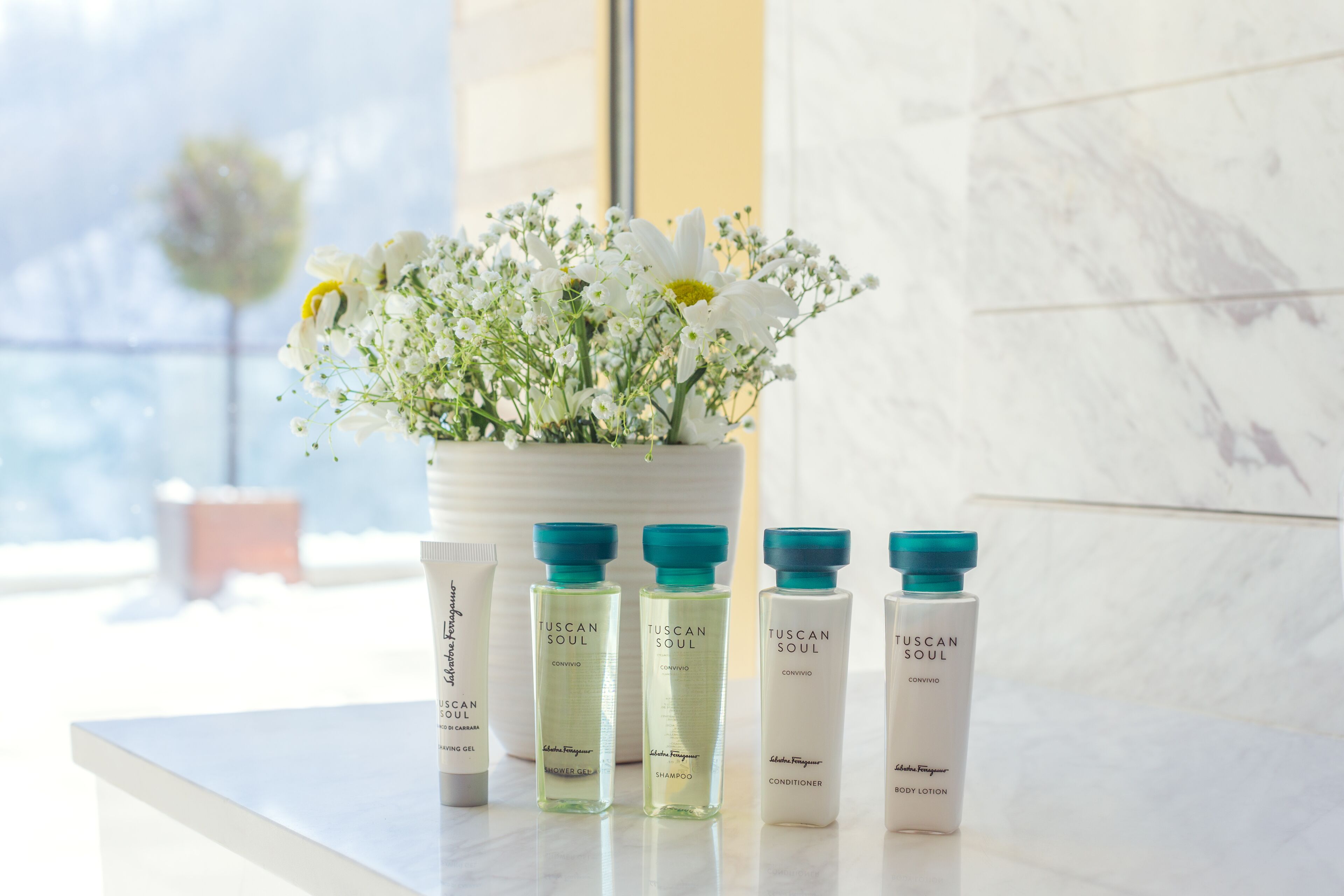 Un meuble-vasque de salle de bain orné d’un vase de fleurs blanches, de quatre produits de la gamme Tuscan Soul et d’un mur en marbre.
