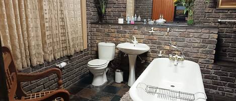 A bathroom with a brick wall, a white toilet, a pedestal sink, and a freestanding bathtub.