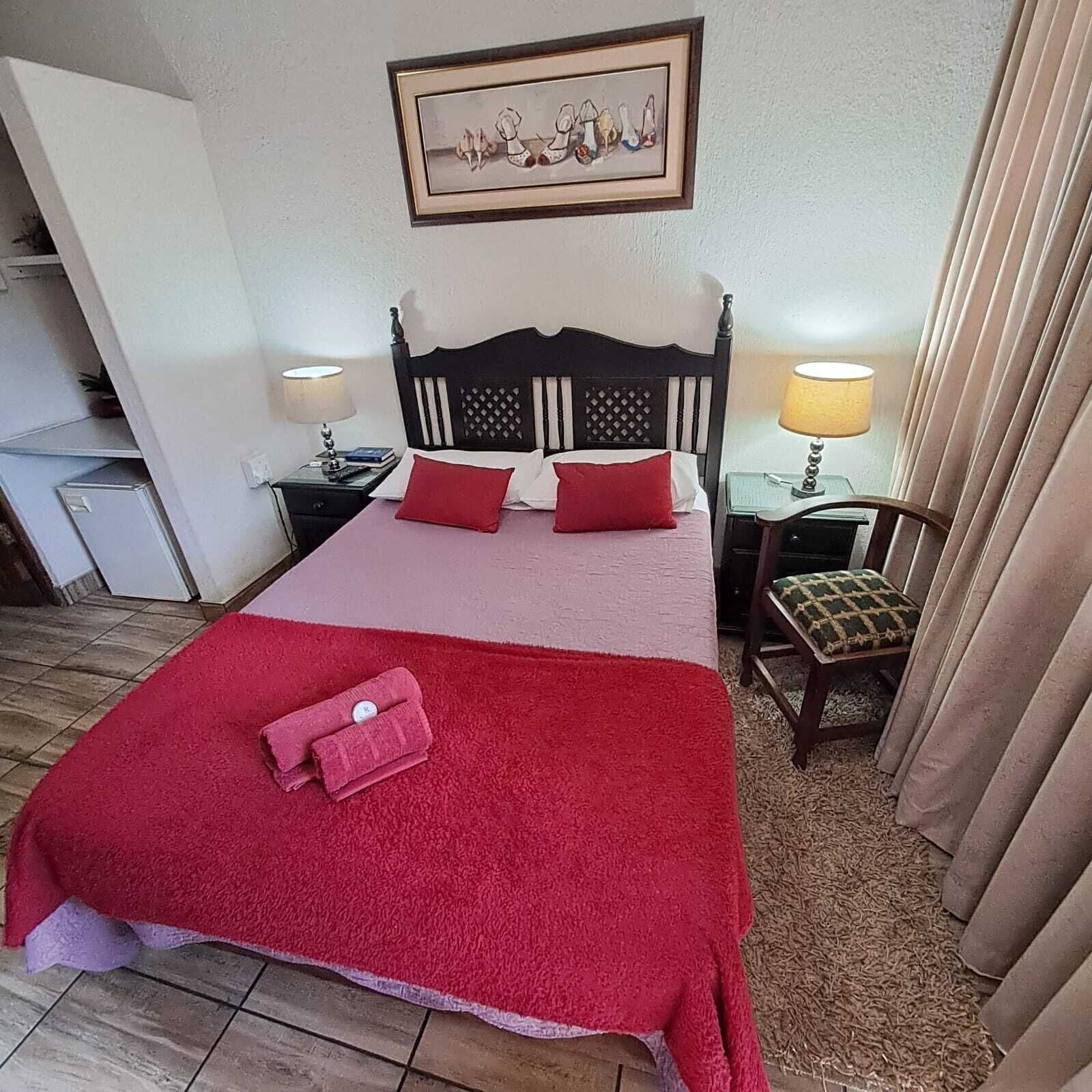 A hotel room with a bed, two bedside tables, a chair, a red blanket, and a framed picture on the wall.