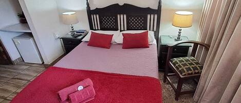 Quarto de hotel com cama, duas mesas de cabeceira, uma cadeira, manta vermelha e um quadro na parede.