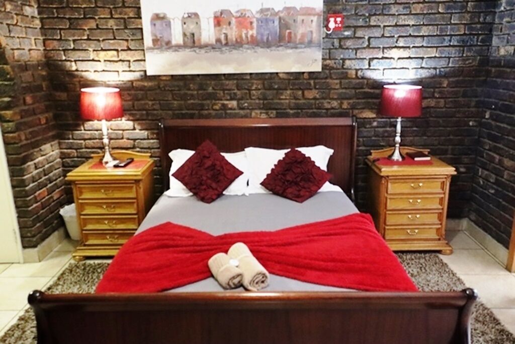 A bedroom with a brick wall, a bed with red bedding, two bedside tables with lamps, and a painting above the bed.