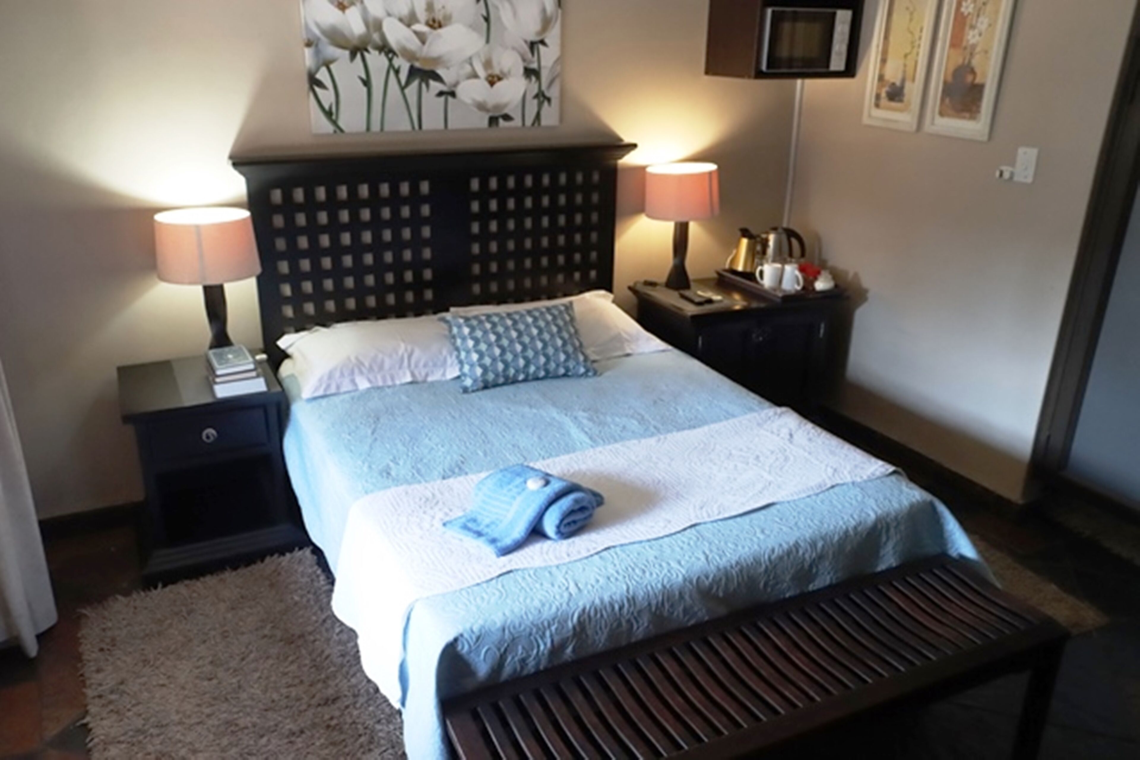 Un lit bien fait, recouvert d’une couvre-lit bleue et blanche, deux lampes de chevet, une table de chevet avec un livre et une petite table, et un téléviseur fixé au mur.