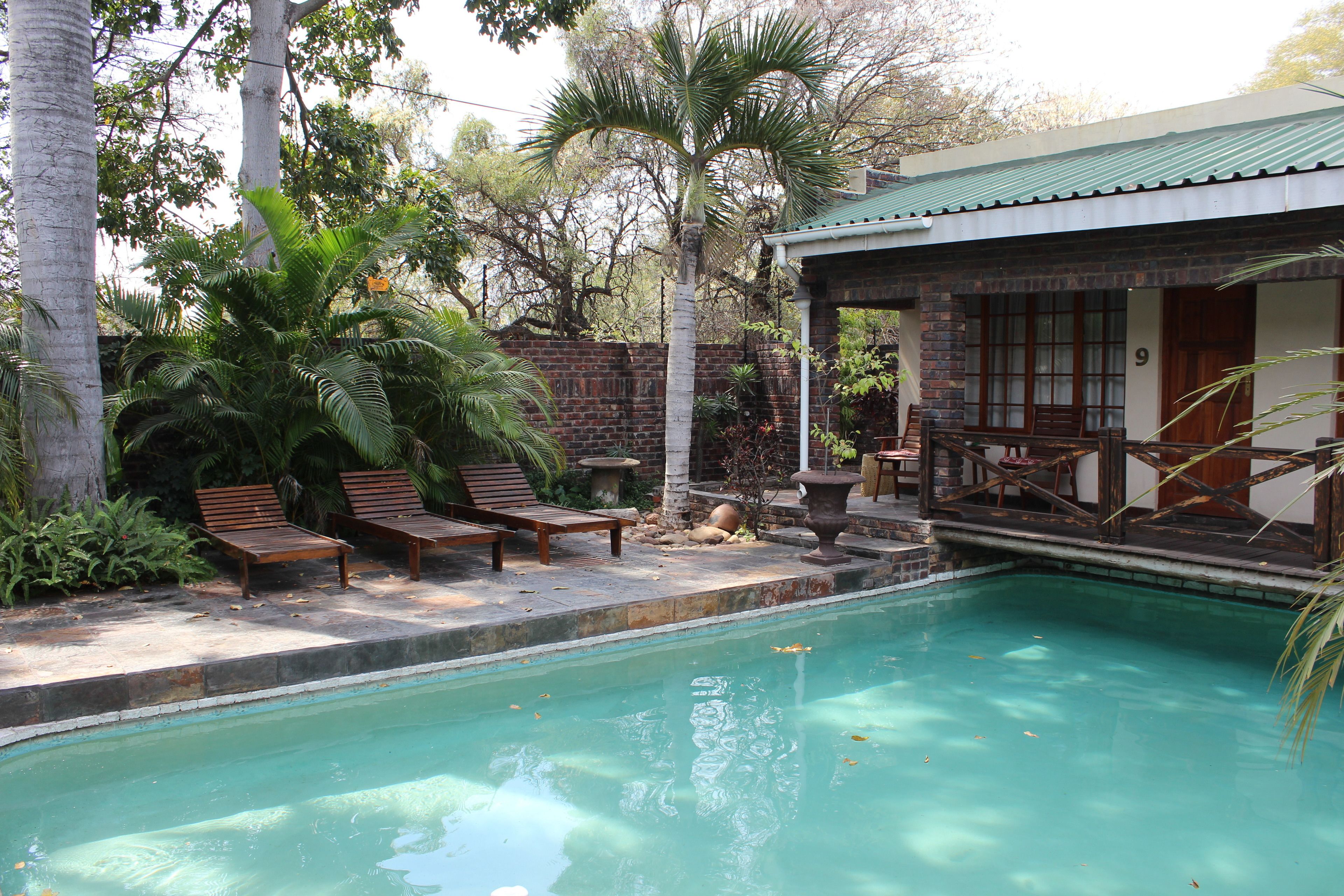 A pool area with wooden lounge chairs, a brick wall, and a building with a green roof.