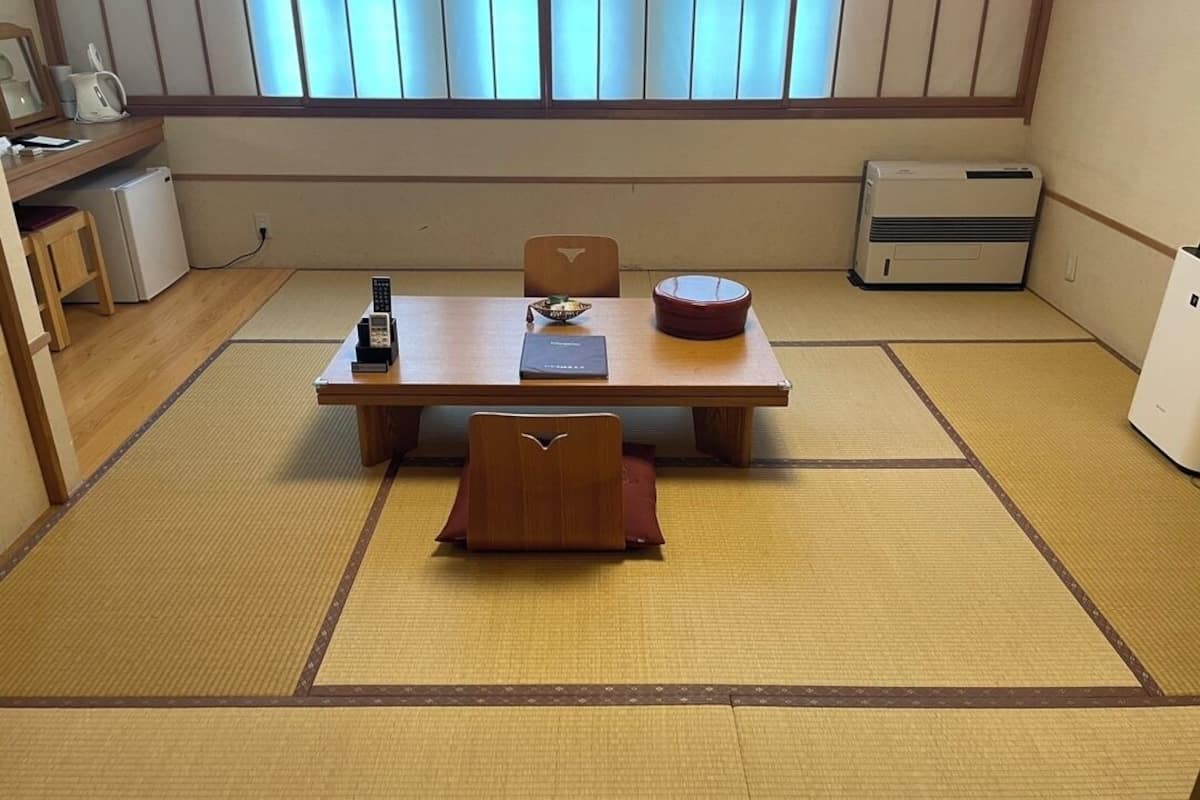 Una tradizionale stanza giapponese con pavimento in tatami, un tavolino con cuscino, un piccolo riscaldatore e una finestra con persiane verticali.