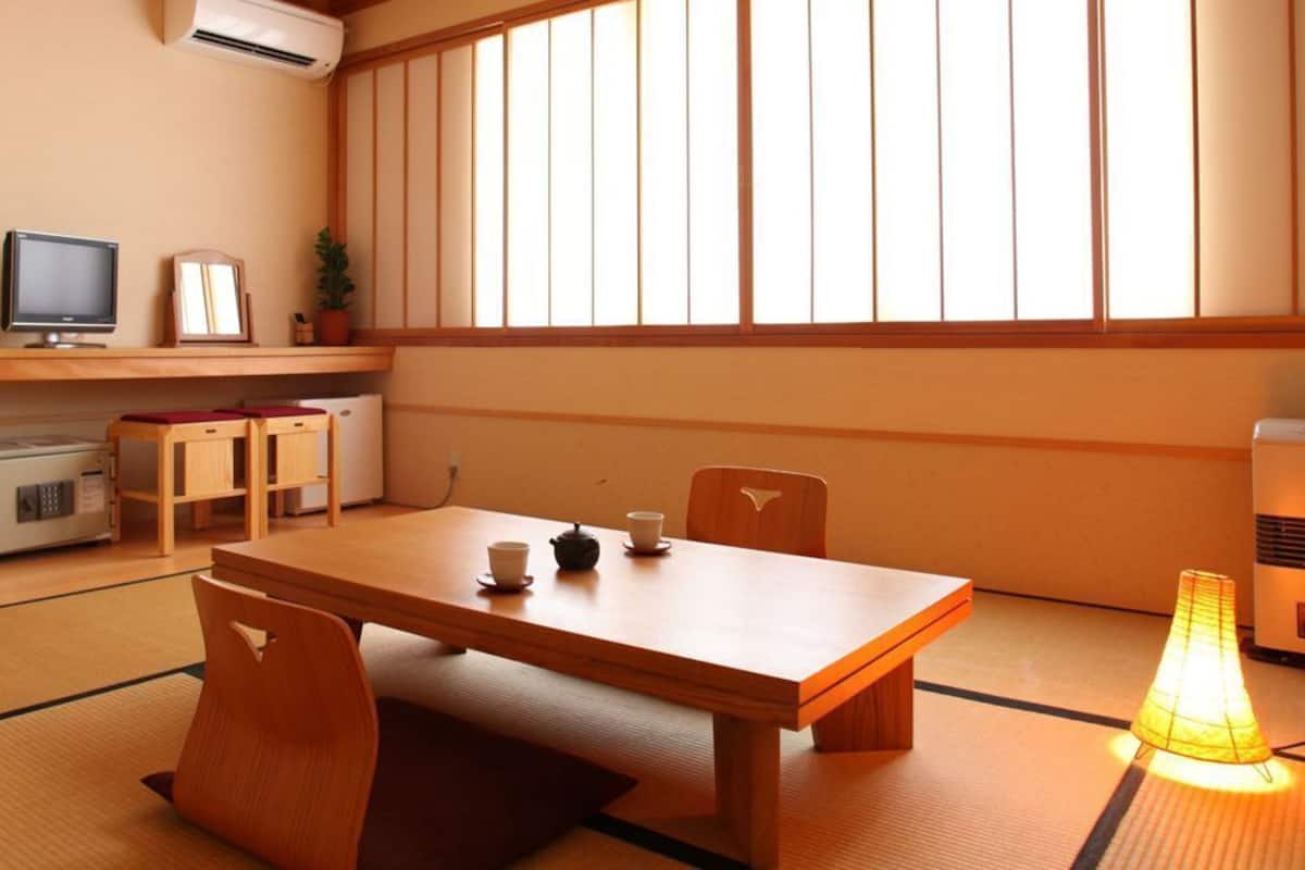 Une pièce de style japonais traditionnel, avec un sol recouvert de tatamis, une table en bois sur laquelle sont posées des tasses et une théière, un téléviseur et un radiateur.