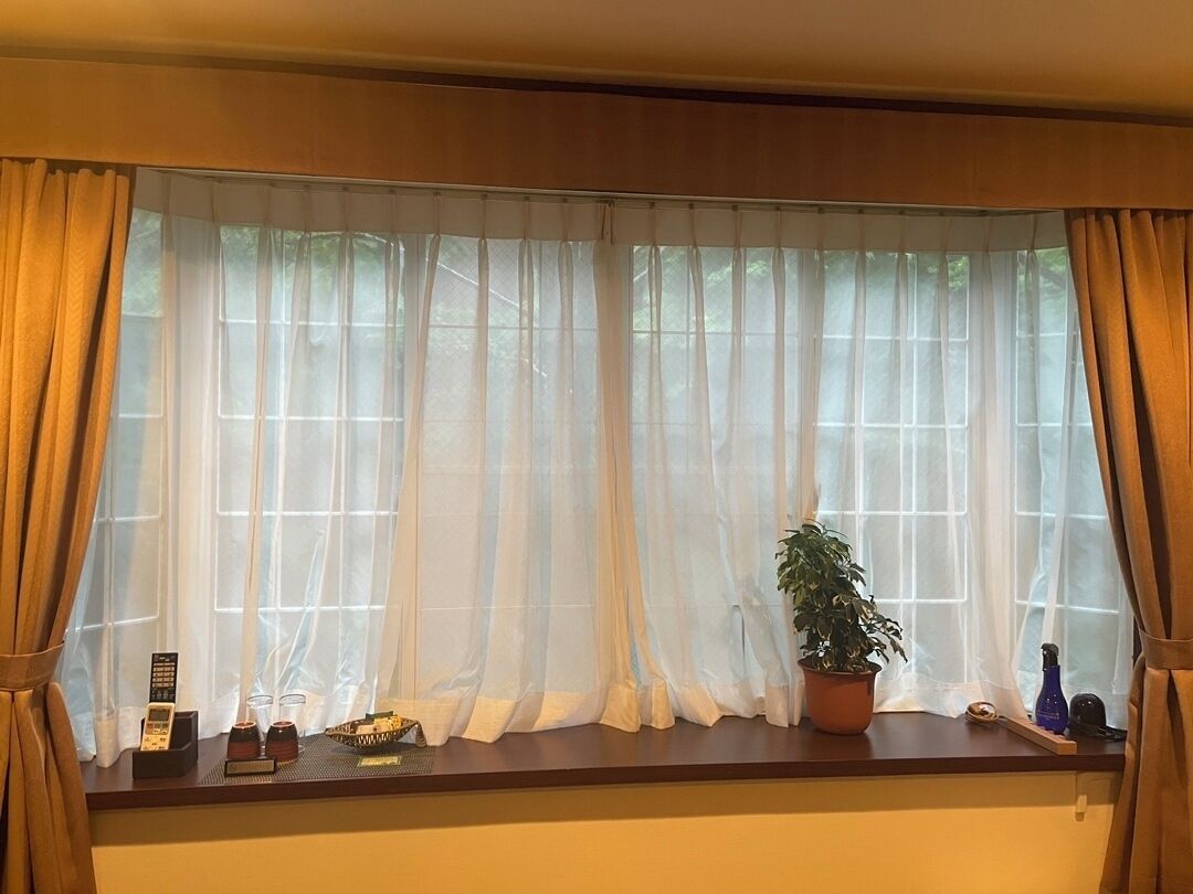 Una ventana con cortinas de encaje, una planta en maceta, una bandeja con objetos y un control remoto en el alféizar.