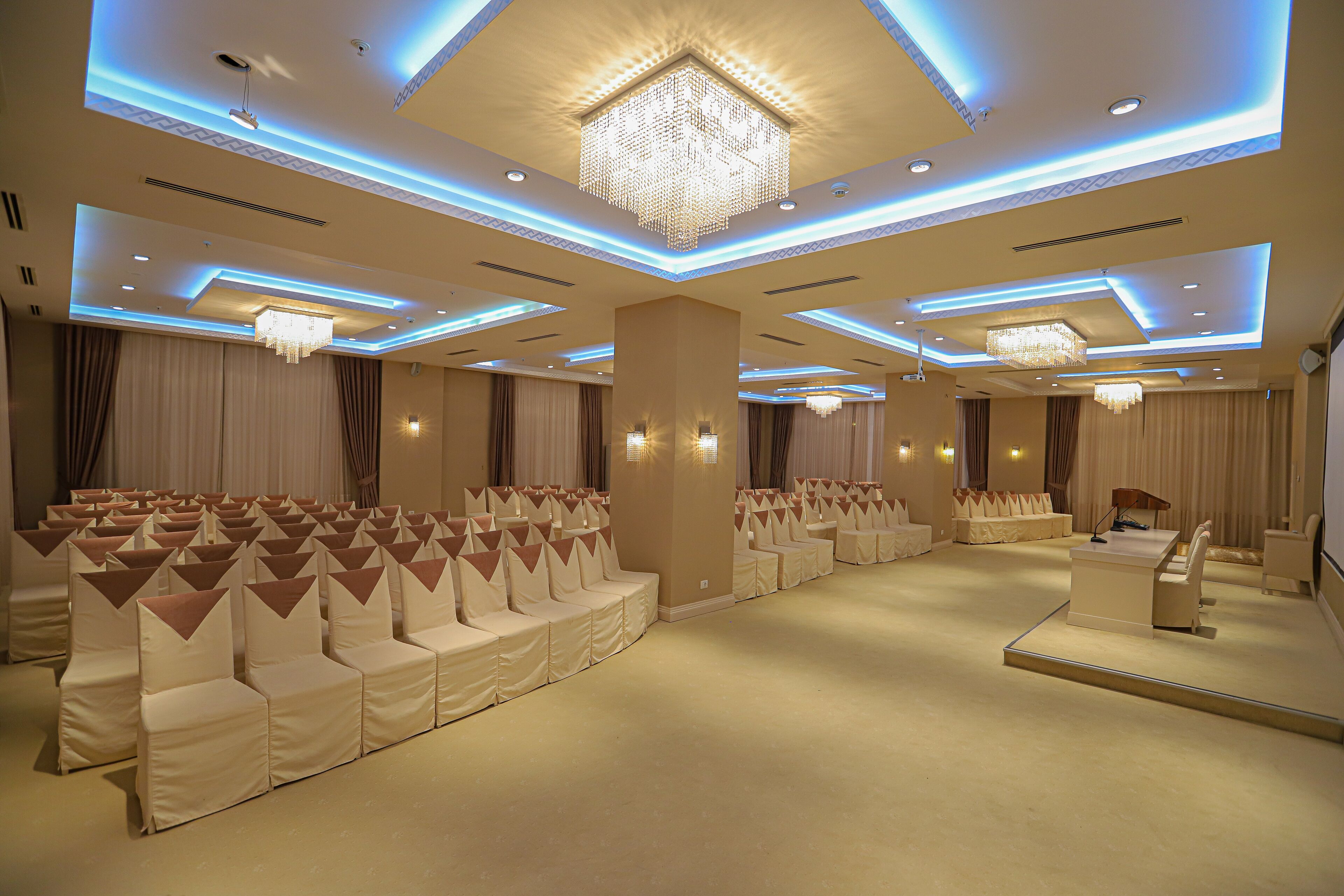 A spacious hall with rows of white chairs, a central stage, and a chandelier.