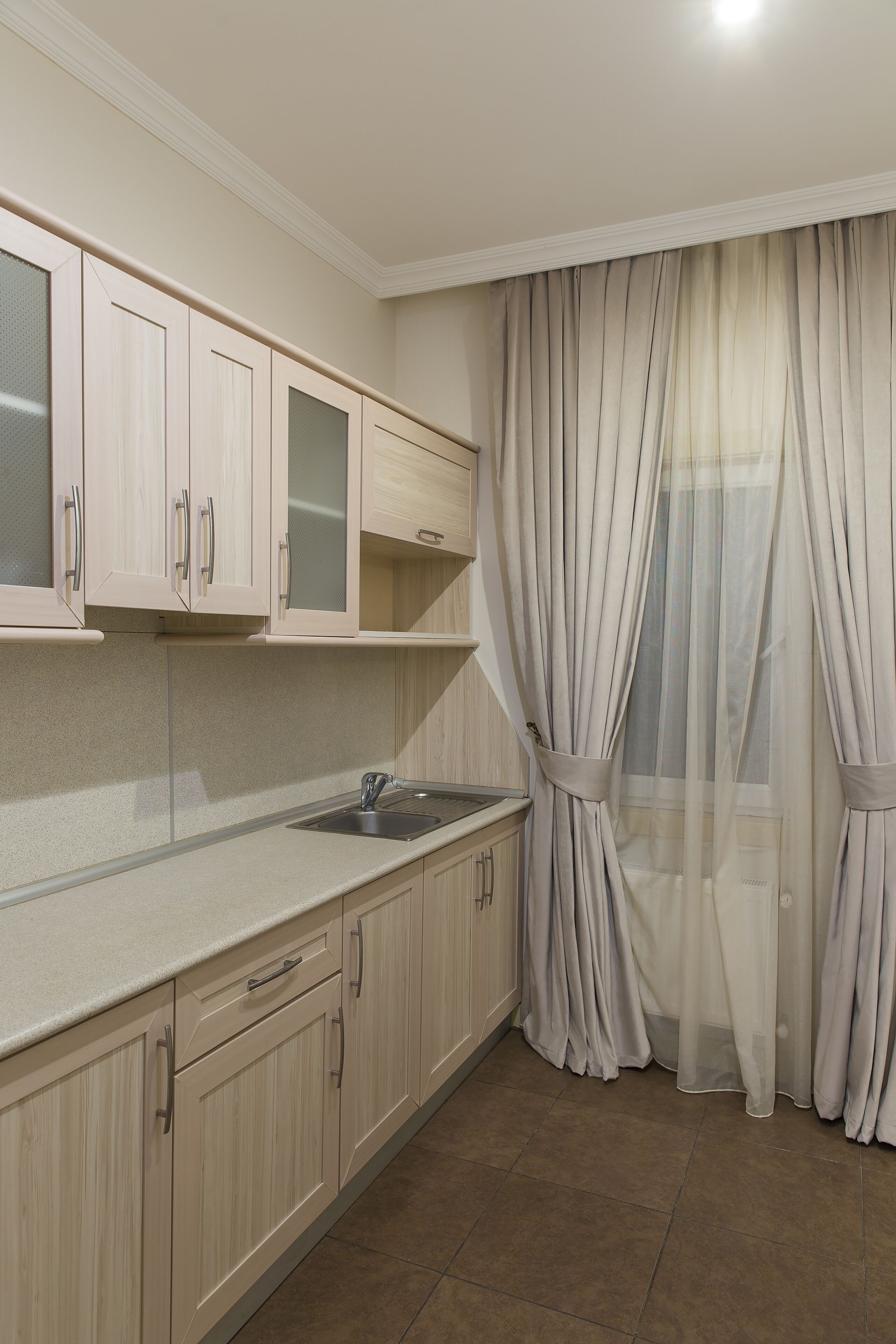 A kitchen with light wood cabinets, a stainless steel sink, and a window with sheer curtains.