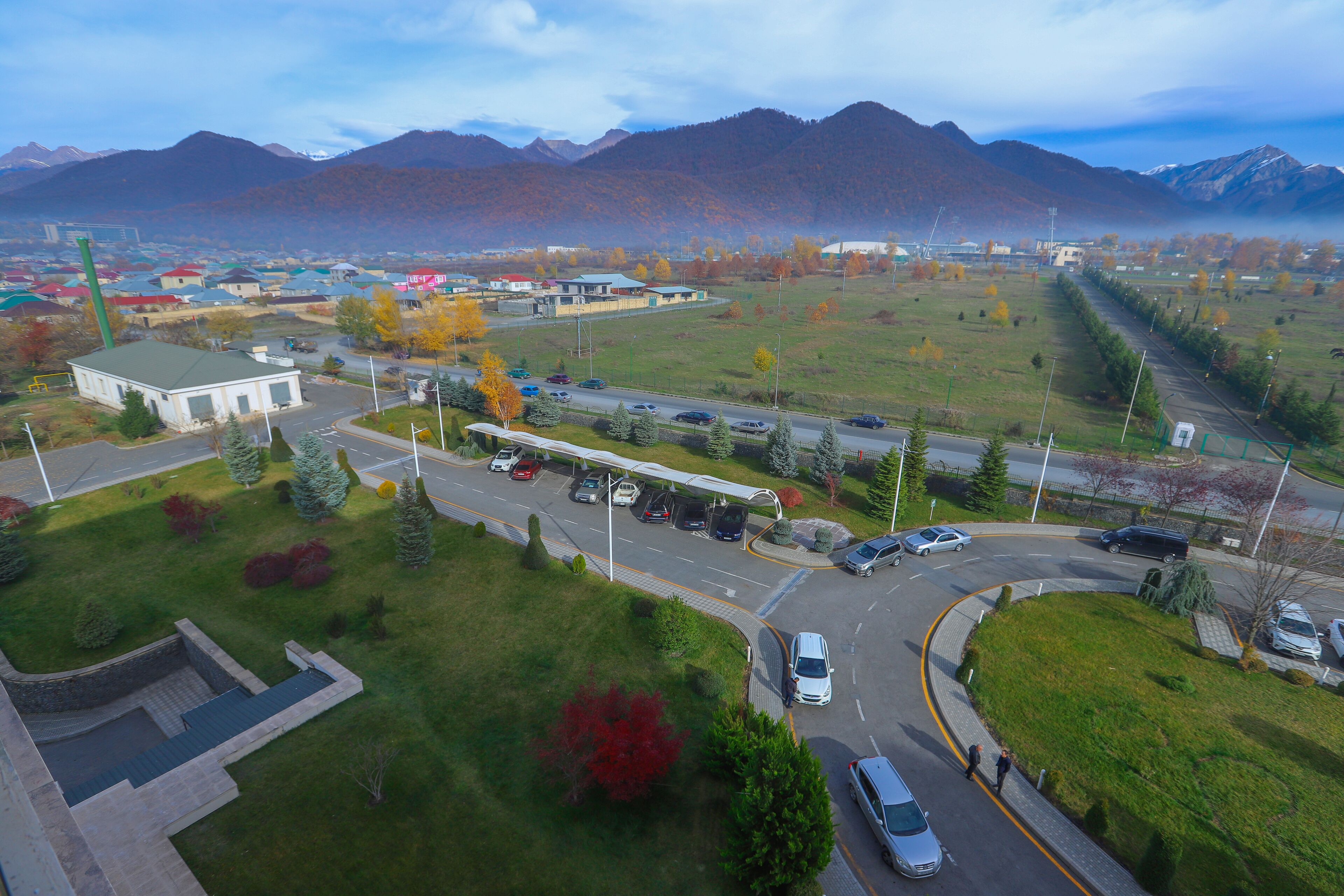 An aerial view of a town with a road leading to a building with a green roof, surrounded by trees and mountains in the background.