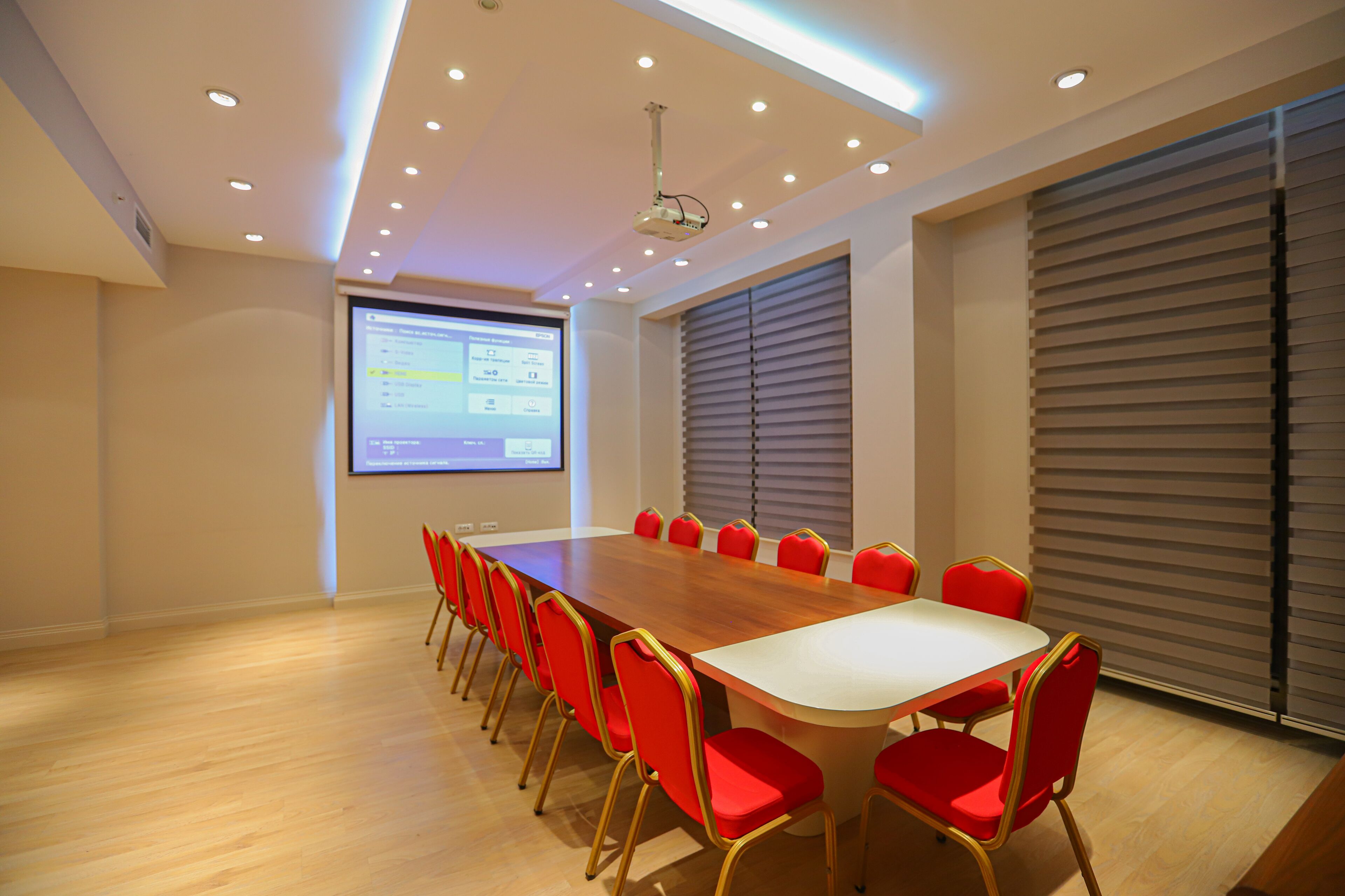 A conference room with a long table, red chairs, a projector screen, and a ceiling with recessed lighting.