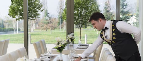 A dining area with a table set for a meal, a waiter attending to the table, and a view of a park through large windows.