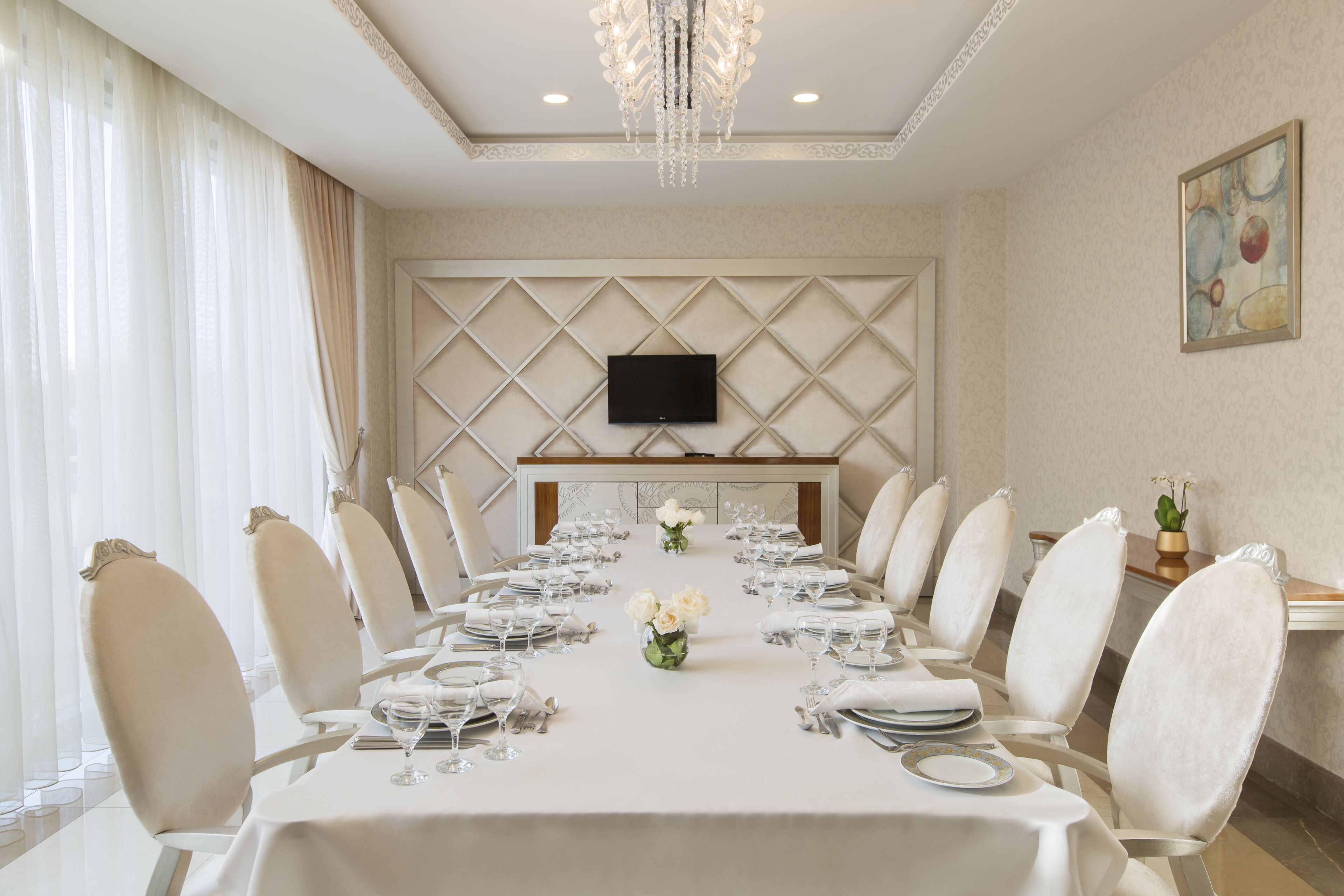 A formal dining room with a long table set for a meal, white chairs, a chandelier, and a flat-screen TV on the wall.