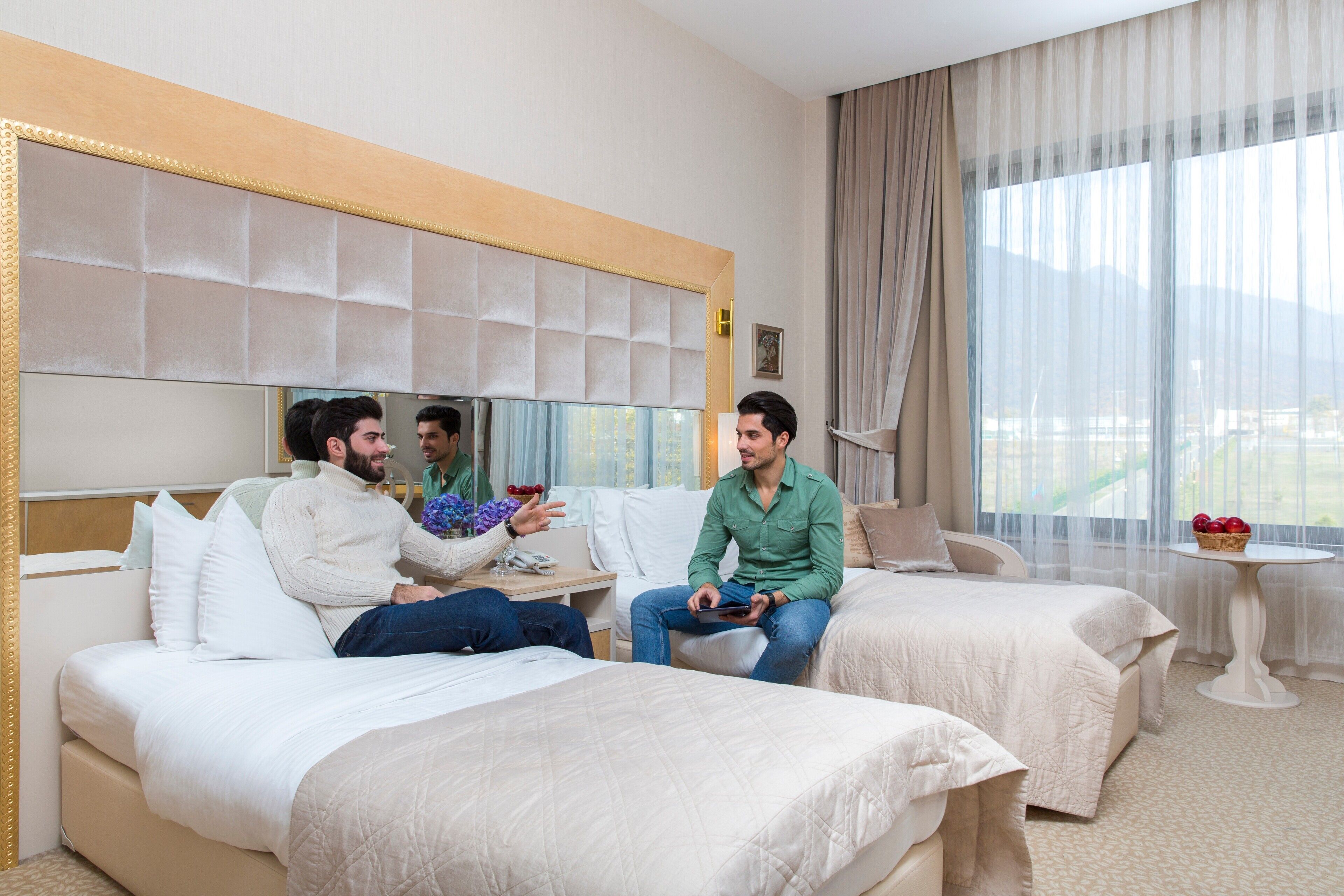 A hotel room with two beds, a large window with curtains, and a small table with a basket of fruit.